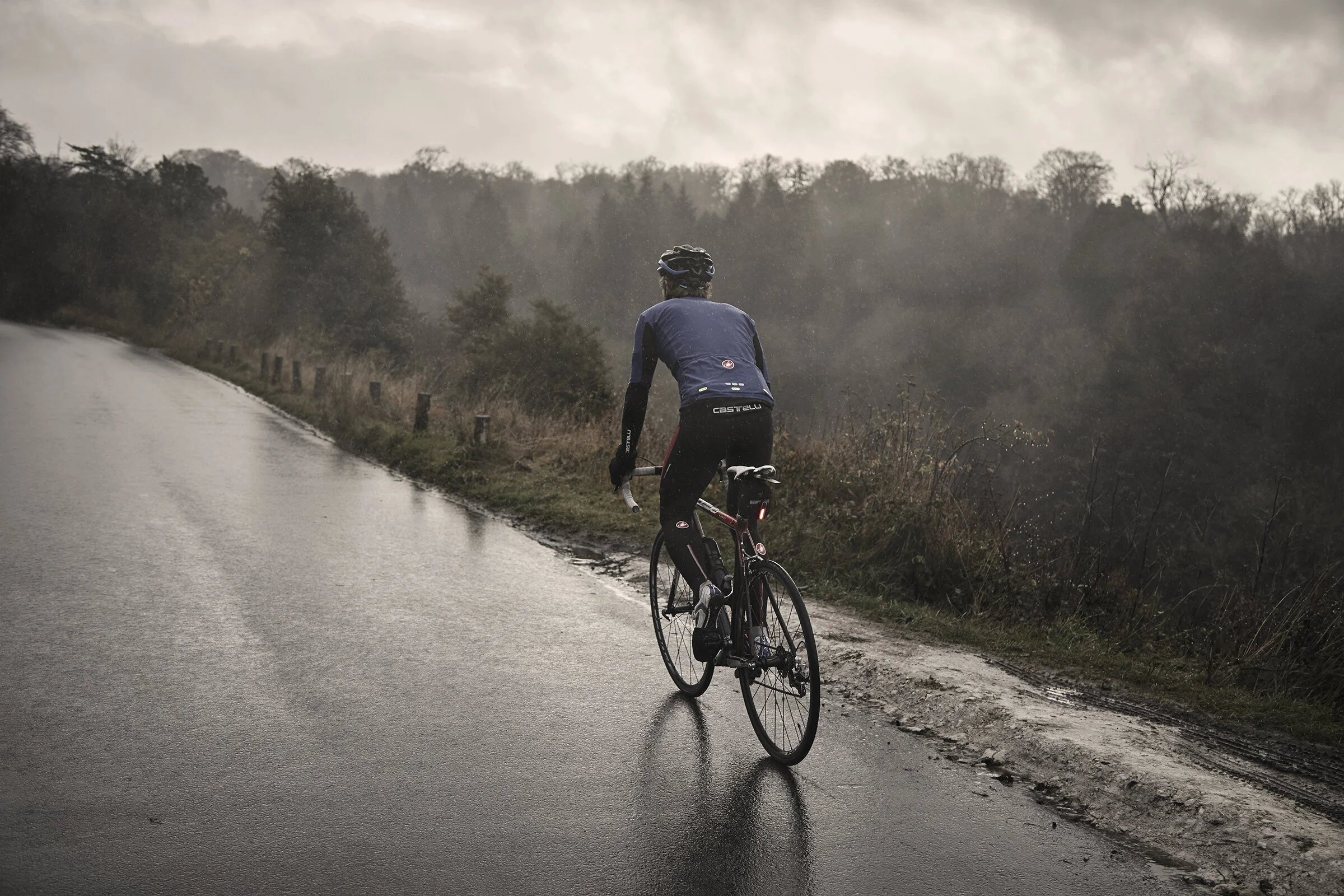 A cyclist riding on a wet, muddy road in a rural area with cloudy, overcast sky and trees in the background.