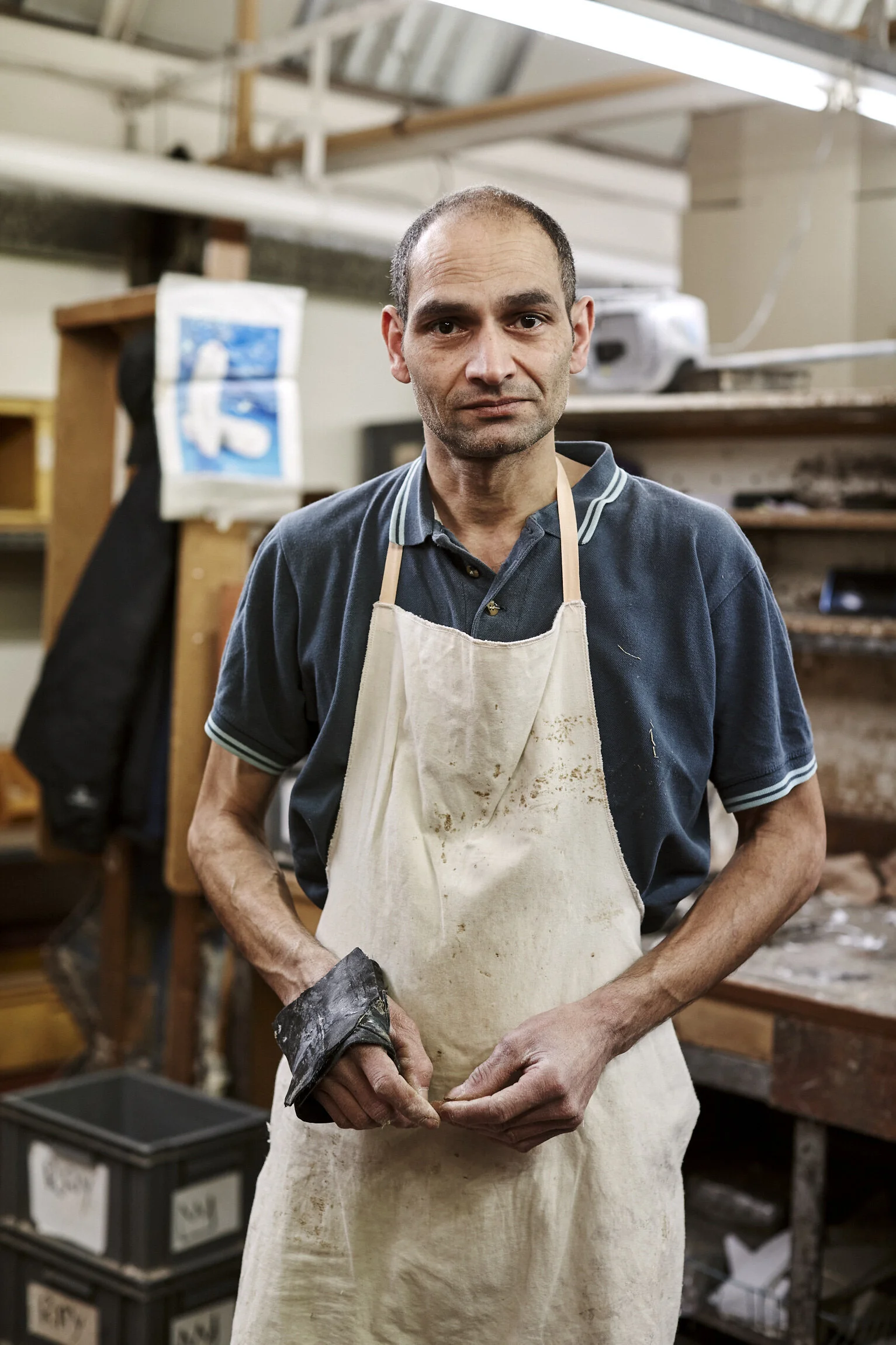 A middle-aged man with short hair wearing a navy blue polo shirt and a stained white apron, standing in a workshop or studio environment, looking directly at the camera with a serious expression.