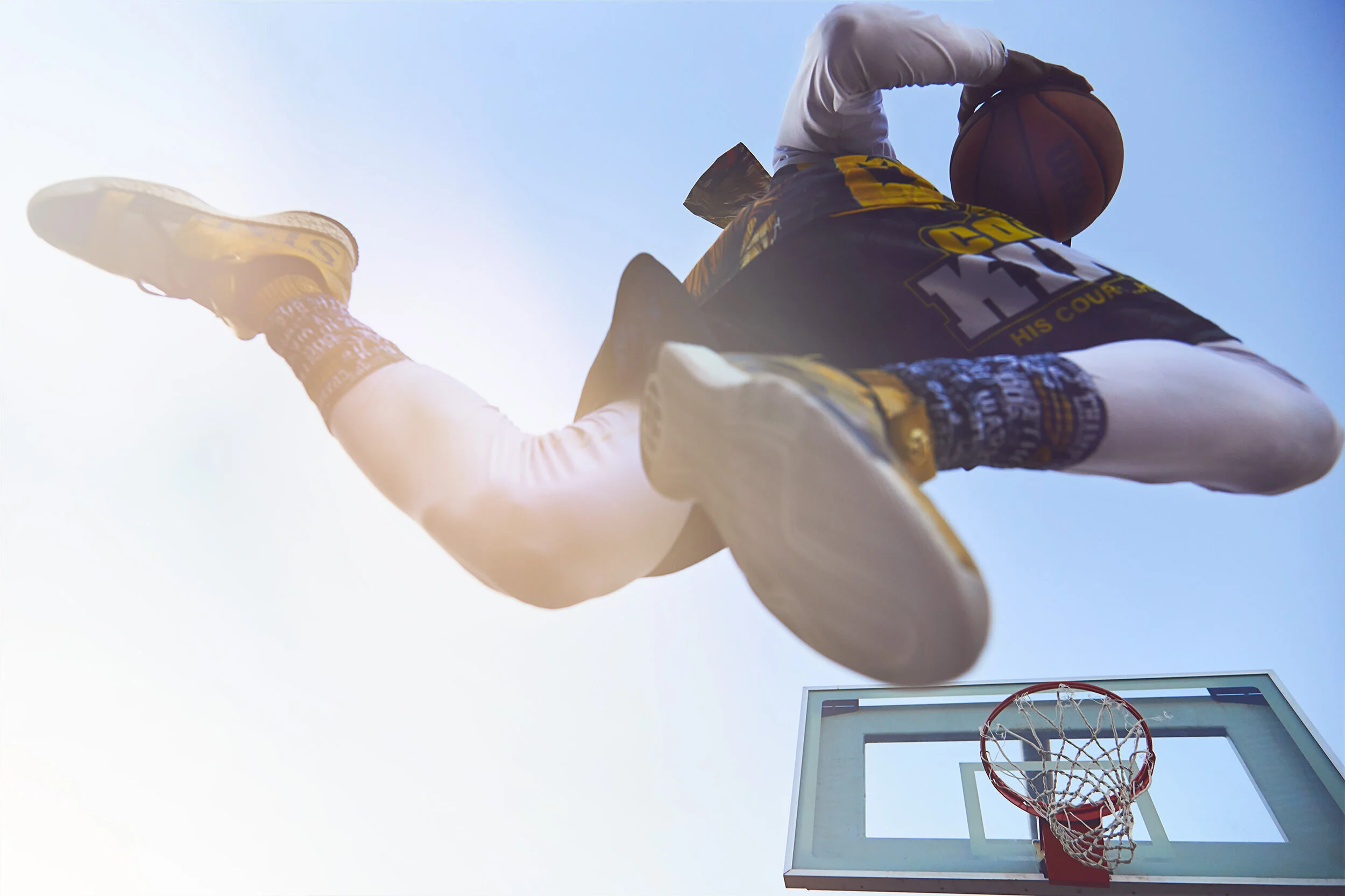 Low-angle view of a young basketball player in a black and yellow jersey with the number 15, airborne, holding a basketball, with a basketball hoop visible below on a sunny day.