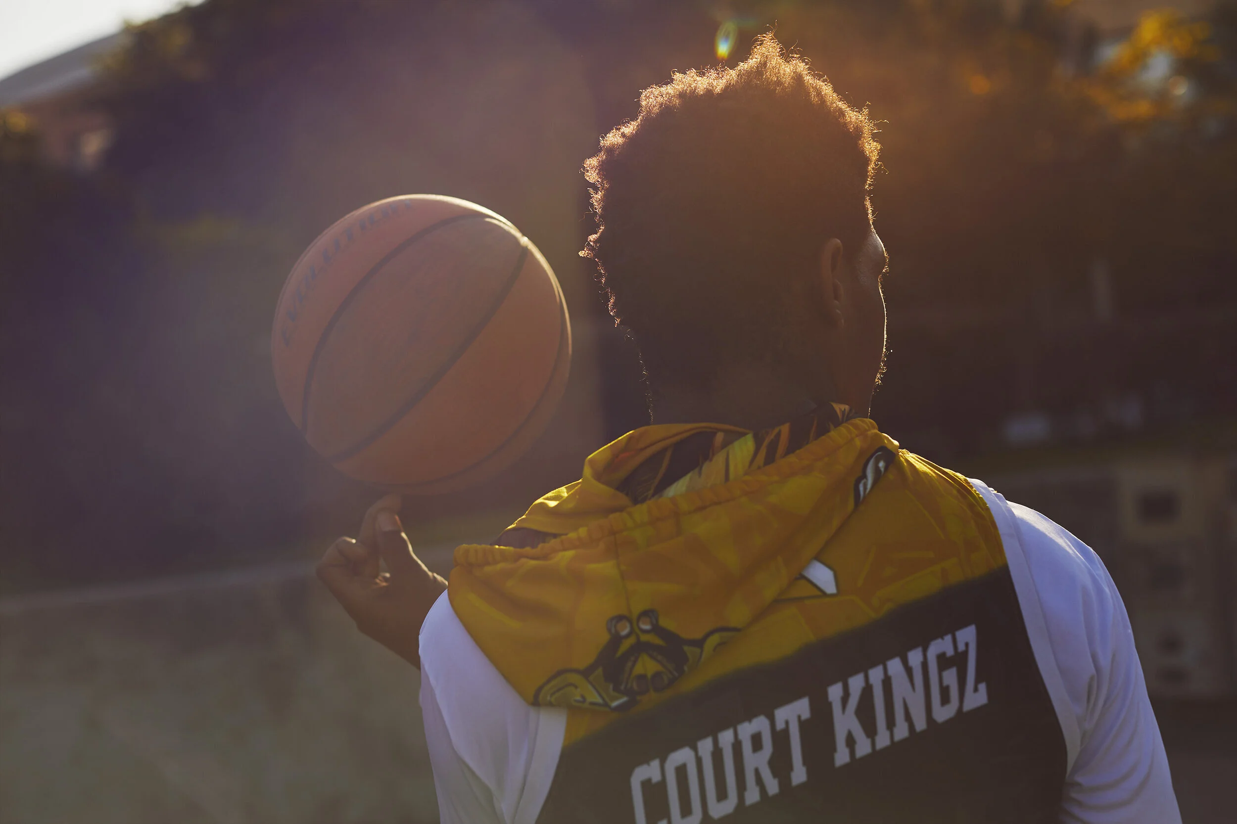 Back view of a young man with curly hair wearing a yellow and black hoodie with ''Court Kingz'' written on it, spinning a basketball on his finger outdoors during sunset.