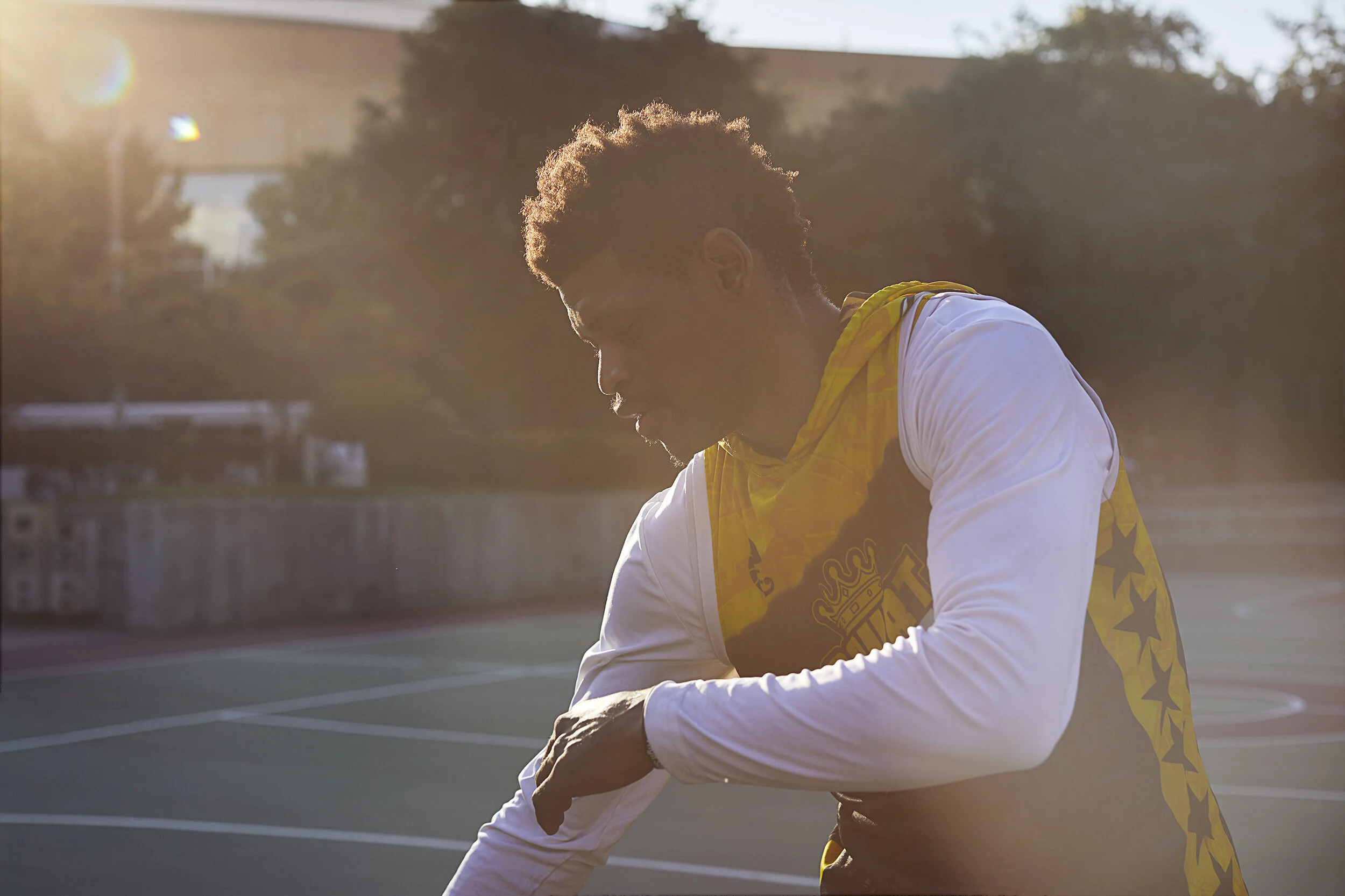 Young man with curly hair and beard wearing a white and yellow sports jersey, adjusting his sleeve on an outdoor basketball court during sunset.