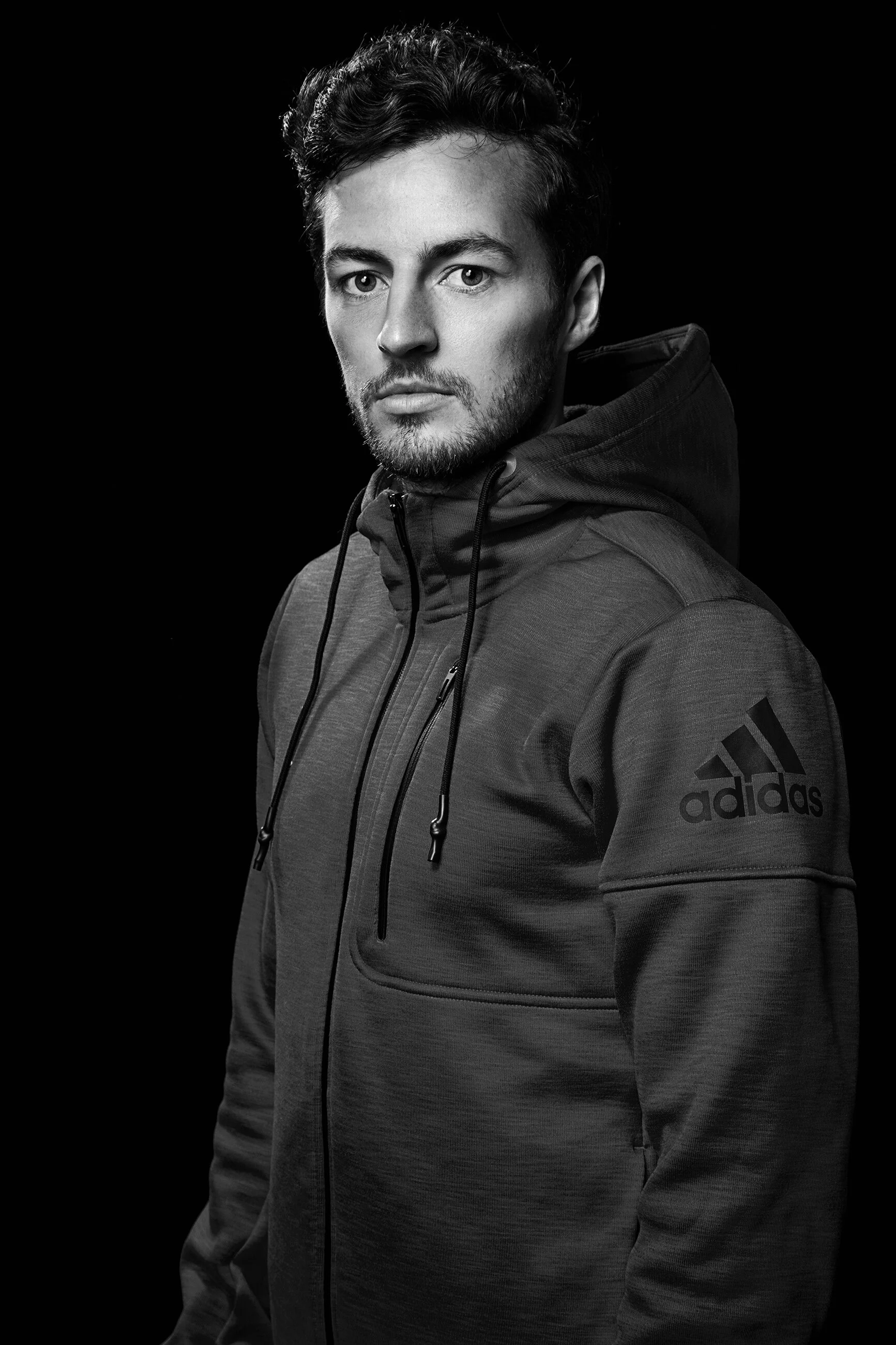 Black and white portrait of a young man wearing a dark Adidas hoodie with logo on the sleeve, looking at the camera with a serious expression.