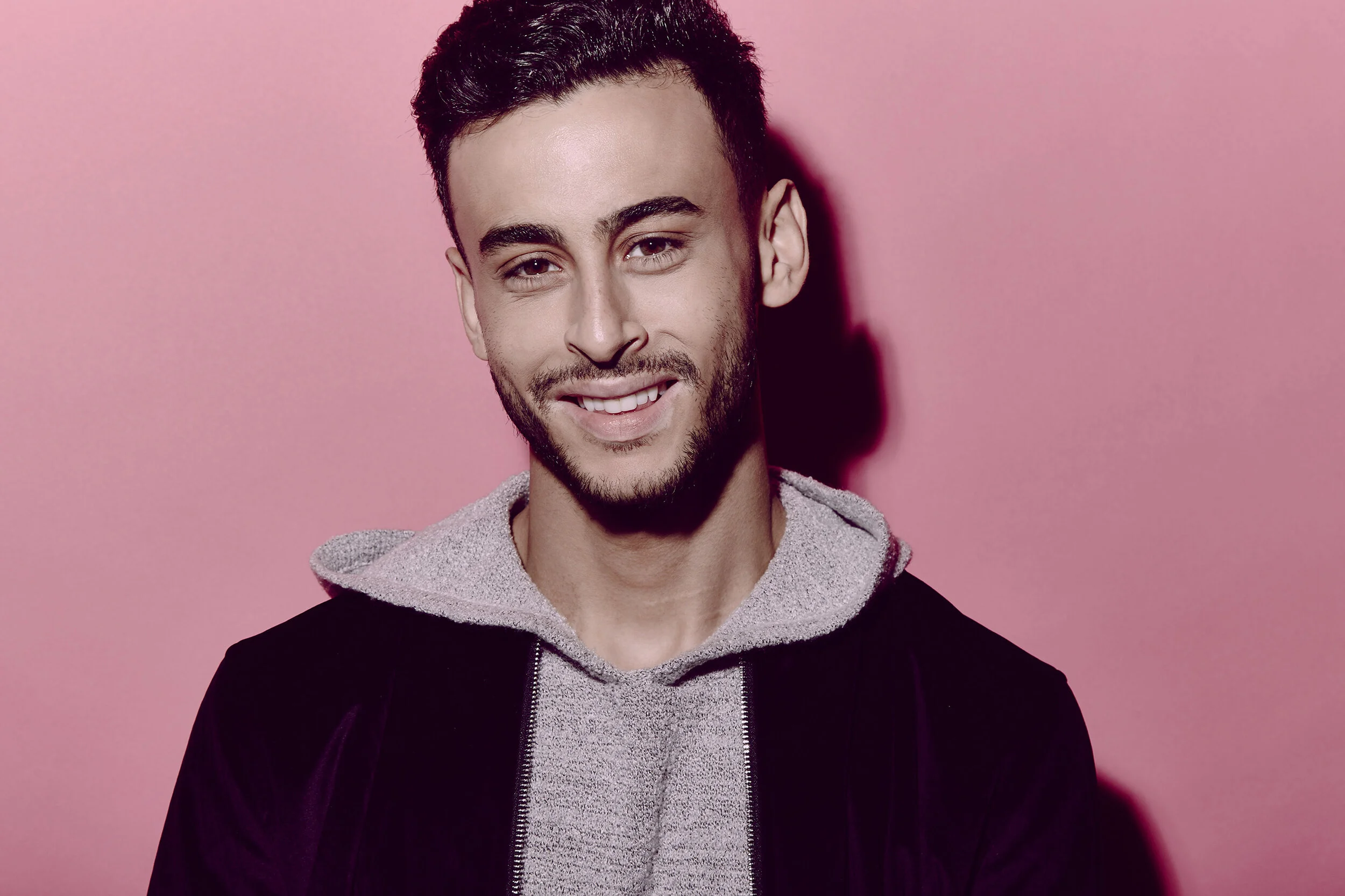 A young man with dark hair, a beard, and a mustache smiling at the camera, wearing a black jacket over a gray hoodie, against a pink background.