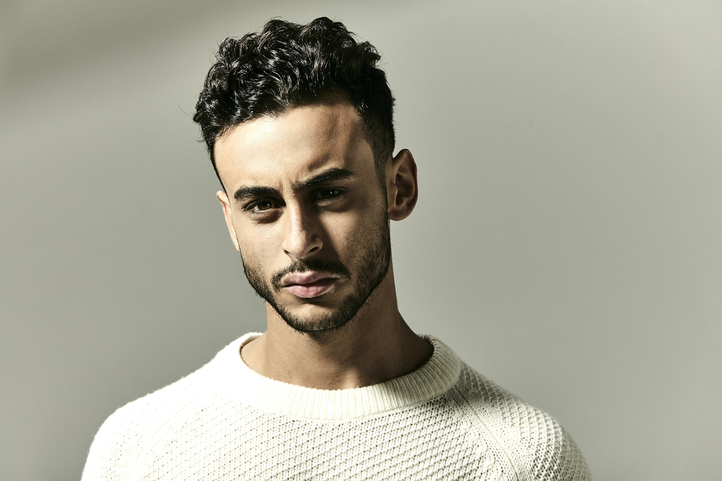 A young man with dark hair and a beard wearing a cream sweater, looking at the camera with a serious expression, standing against a plain neutral background.
