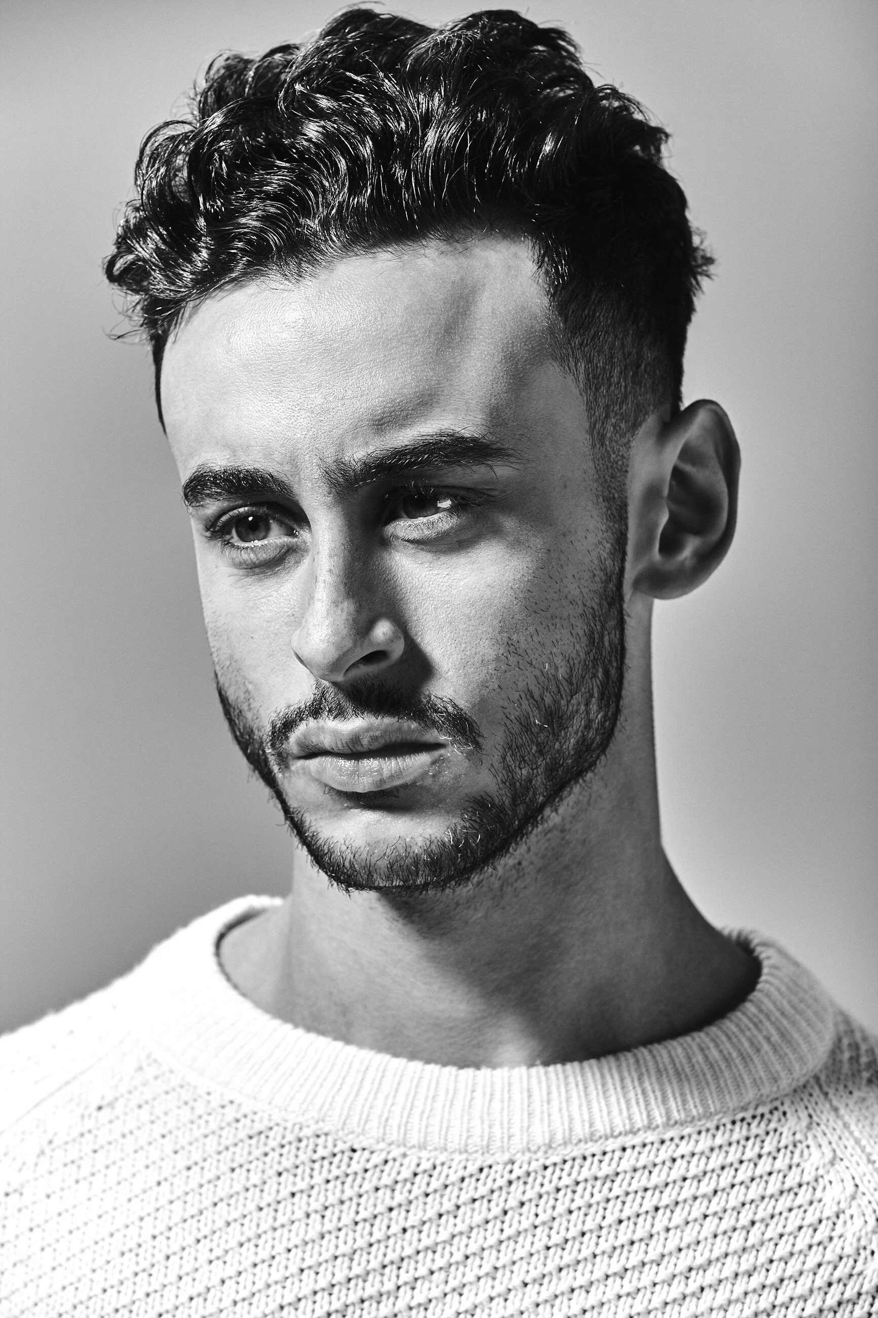 Black and white portrait of a young man with short, curly hair and a beard, wearing a textured sweater, looking pensively to the side.
