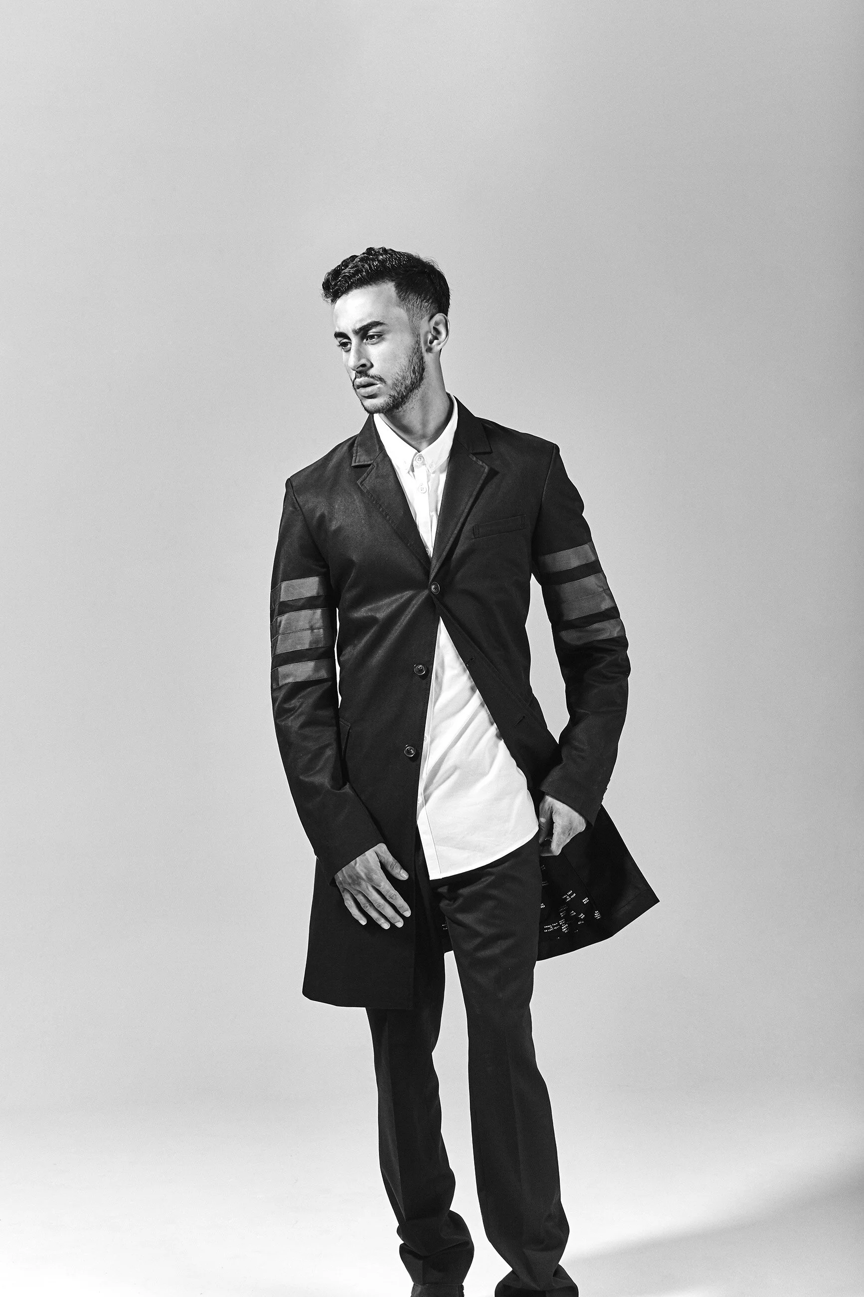 A young man with styled hair and a beard wearing a black coat with striped sleeves, a white shirt, and black pants. He stands against a plain, light gray background, looking off to the side with a serious expression.