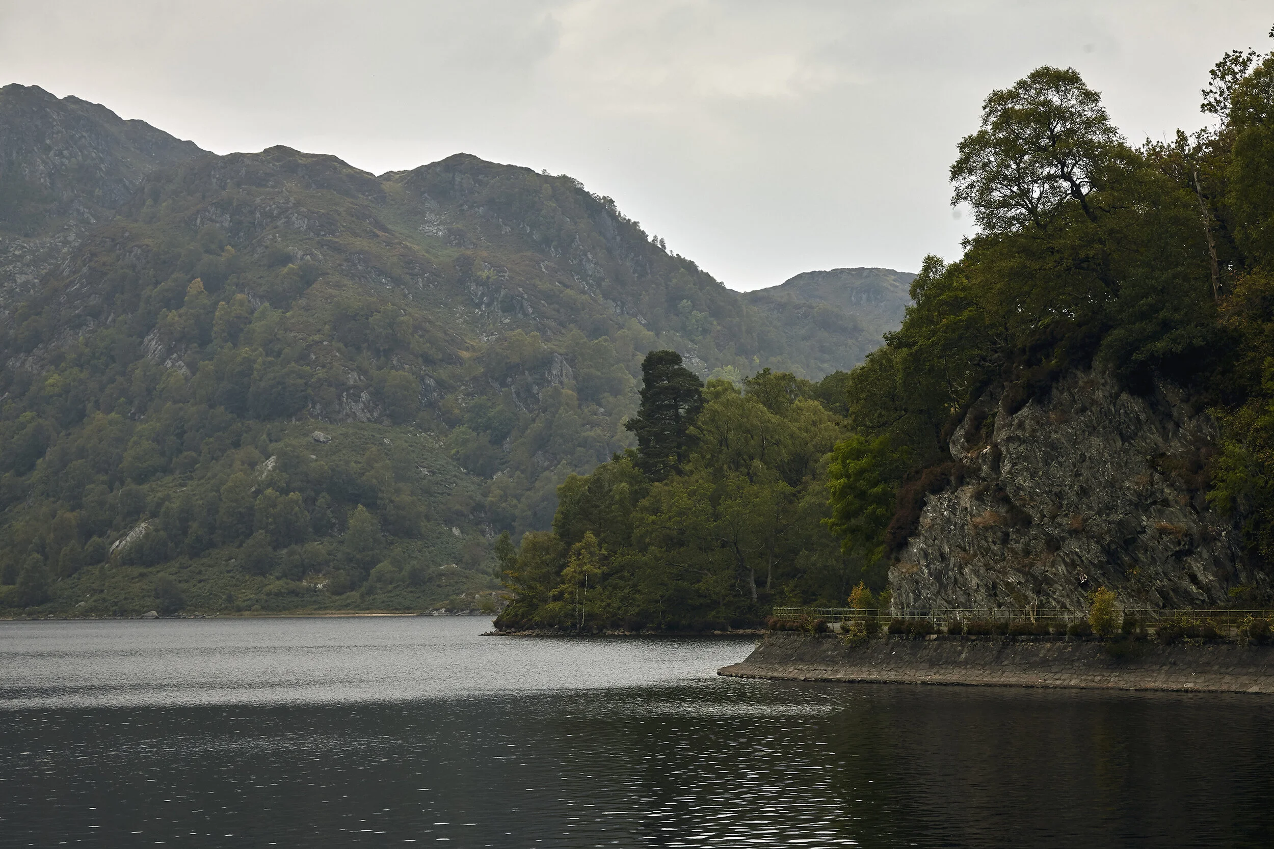 A body of water surrounded by forested hills and mountains under an overcast sky.