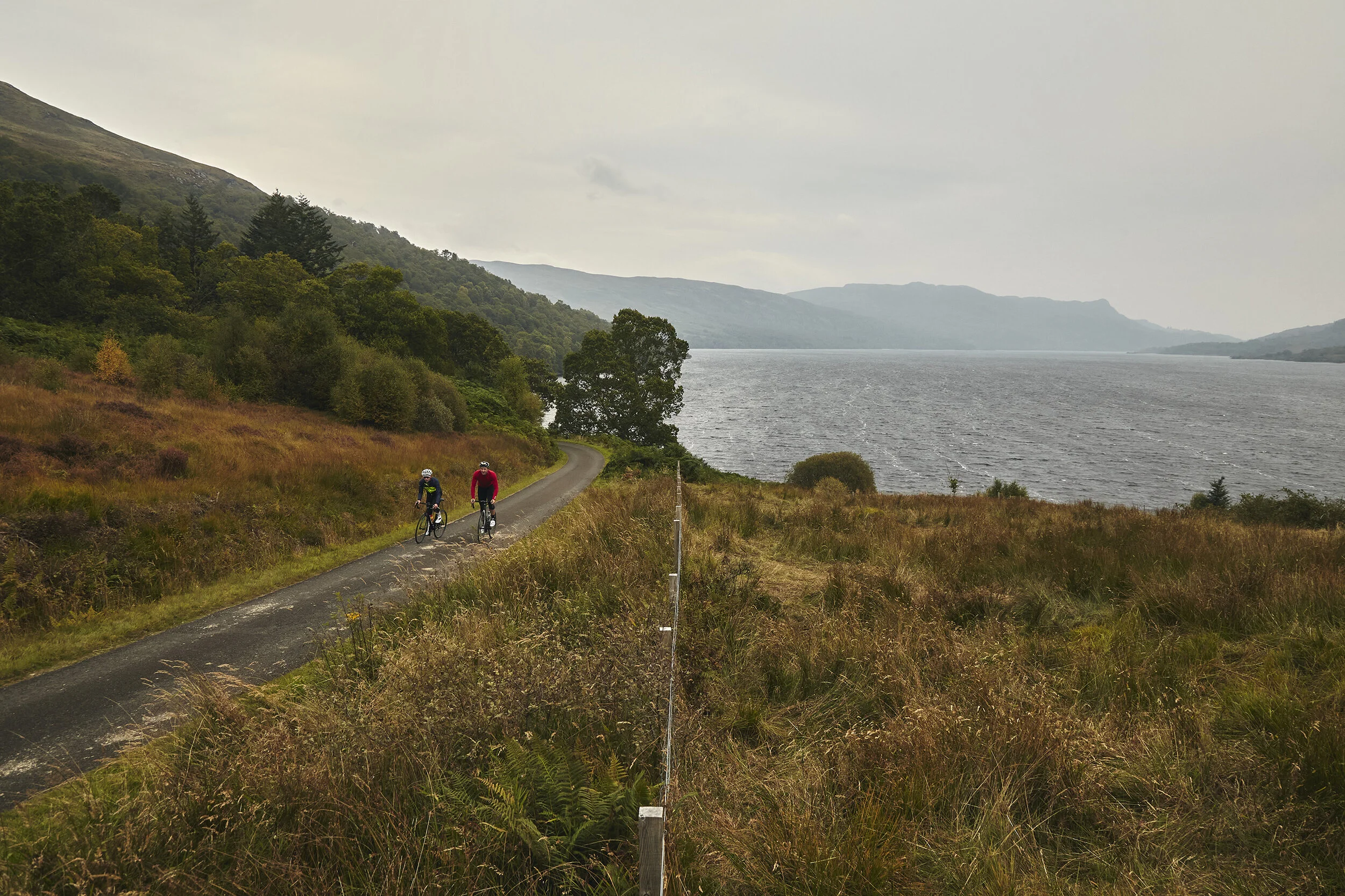 Two cyclists on a winding path near a lake, surrounded by grassy and wooded hills under an overcast sky.