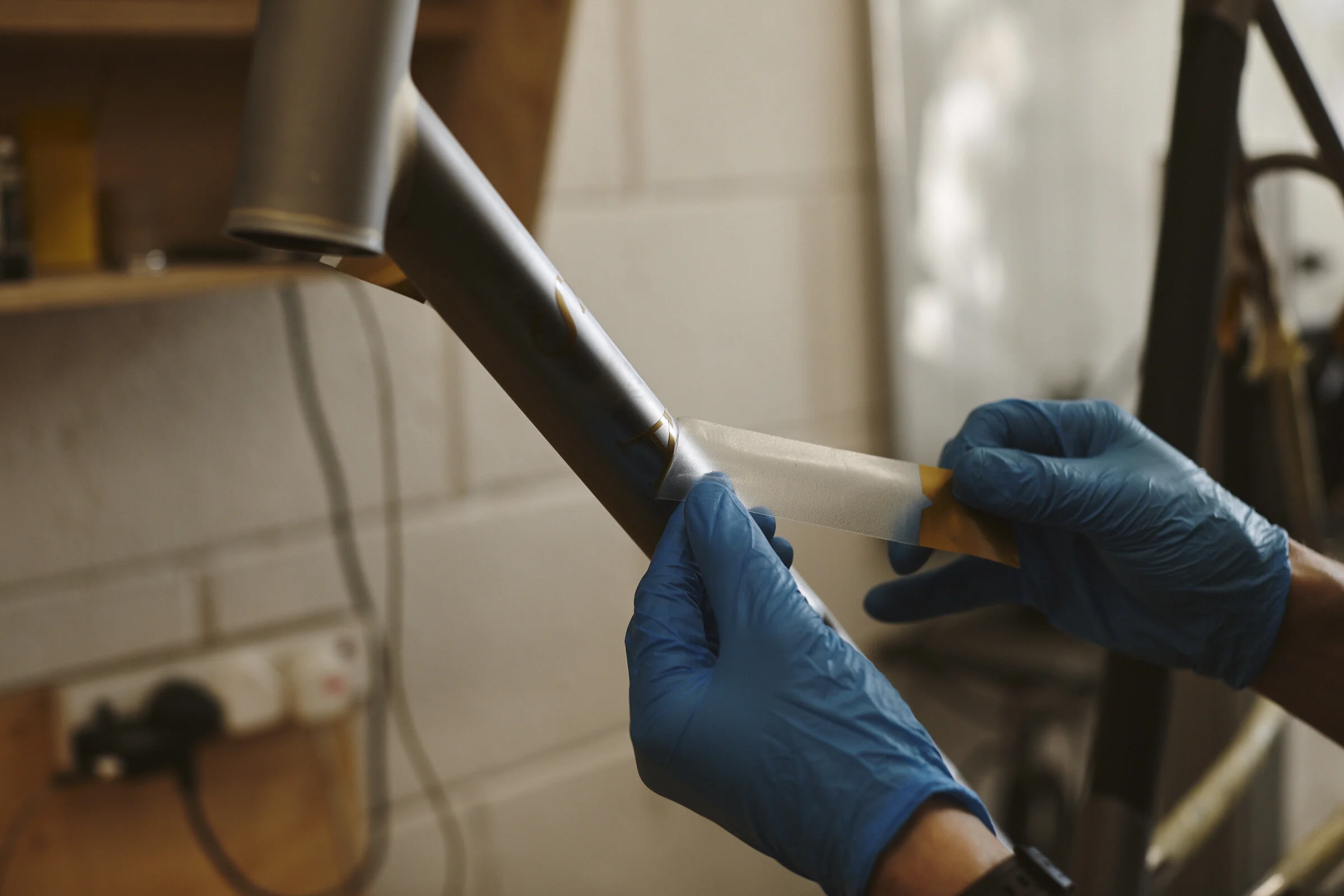 Hands wearing blue gloves applying protective tape to a metallic bicycle frame in a workshop.