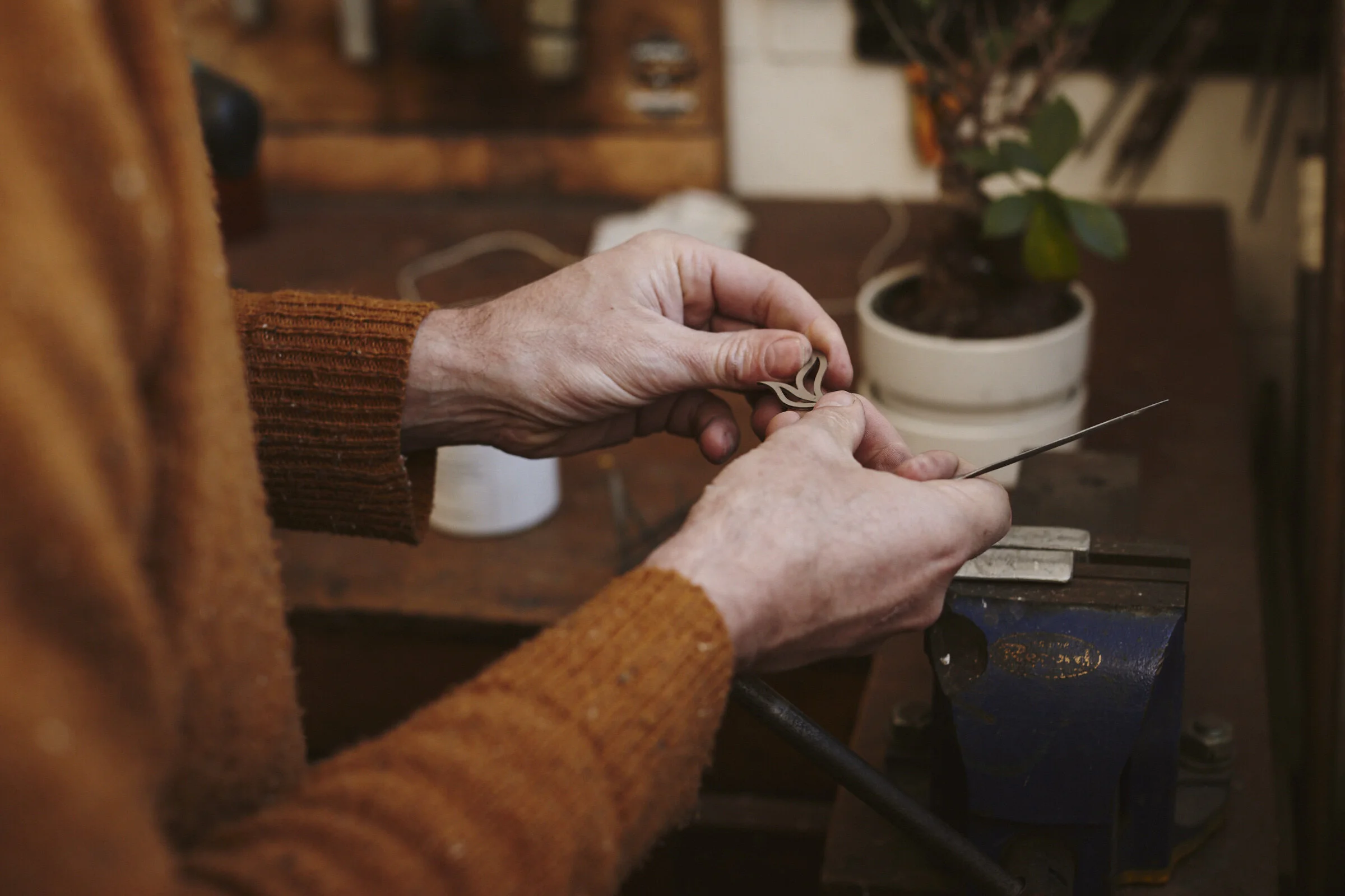 A person wearing an orange sweater working on a jewelry piece in a workshop, with a potted plant in the background.