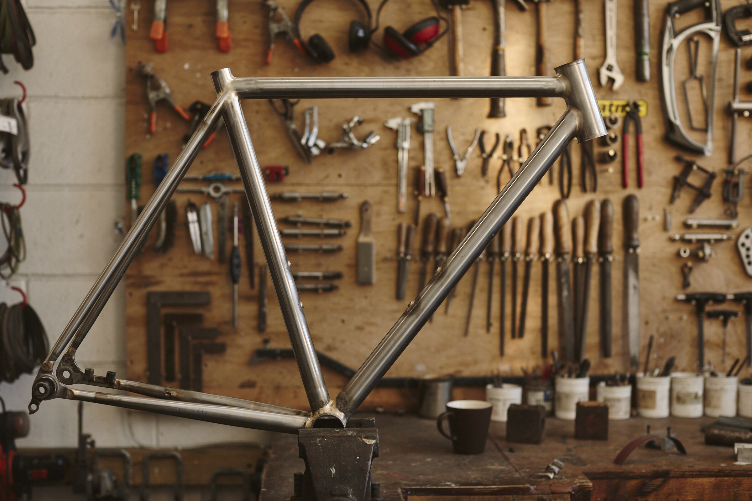 A bicycle frame made of metal in a workshop with tools hanging on a wooden wall in the background.