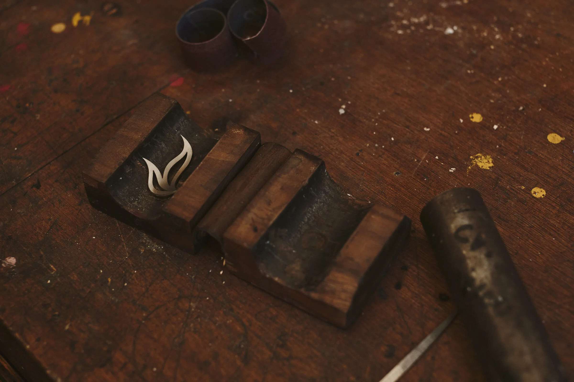 A wooden work surface with a rubber stamp and two rings, with a tattooed tool nearby, surrounded by paint splatters and small debris.