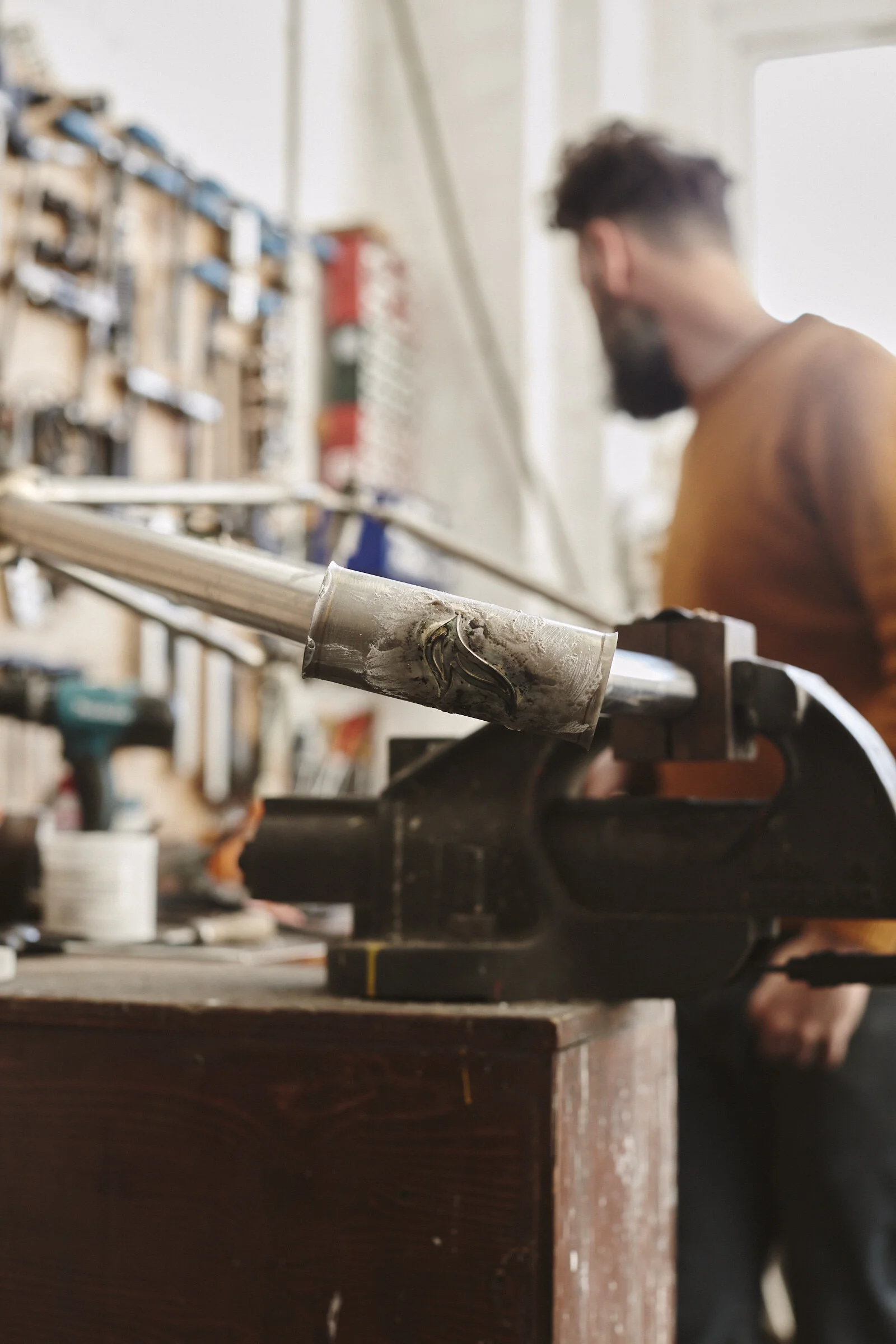 A man working in a workshop, focused on a metal pipe secured in a vise, with tools and equipment on a wooden workbench and a pegboard with tools in the background.