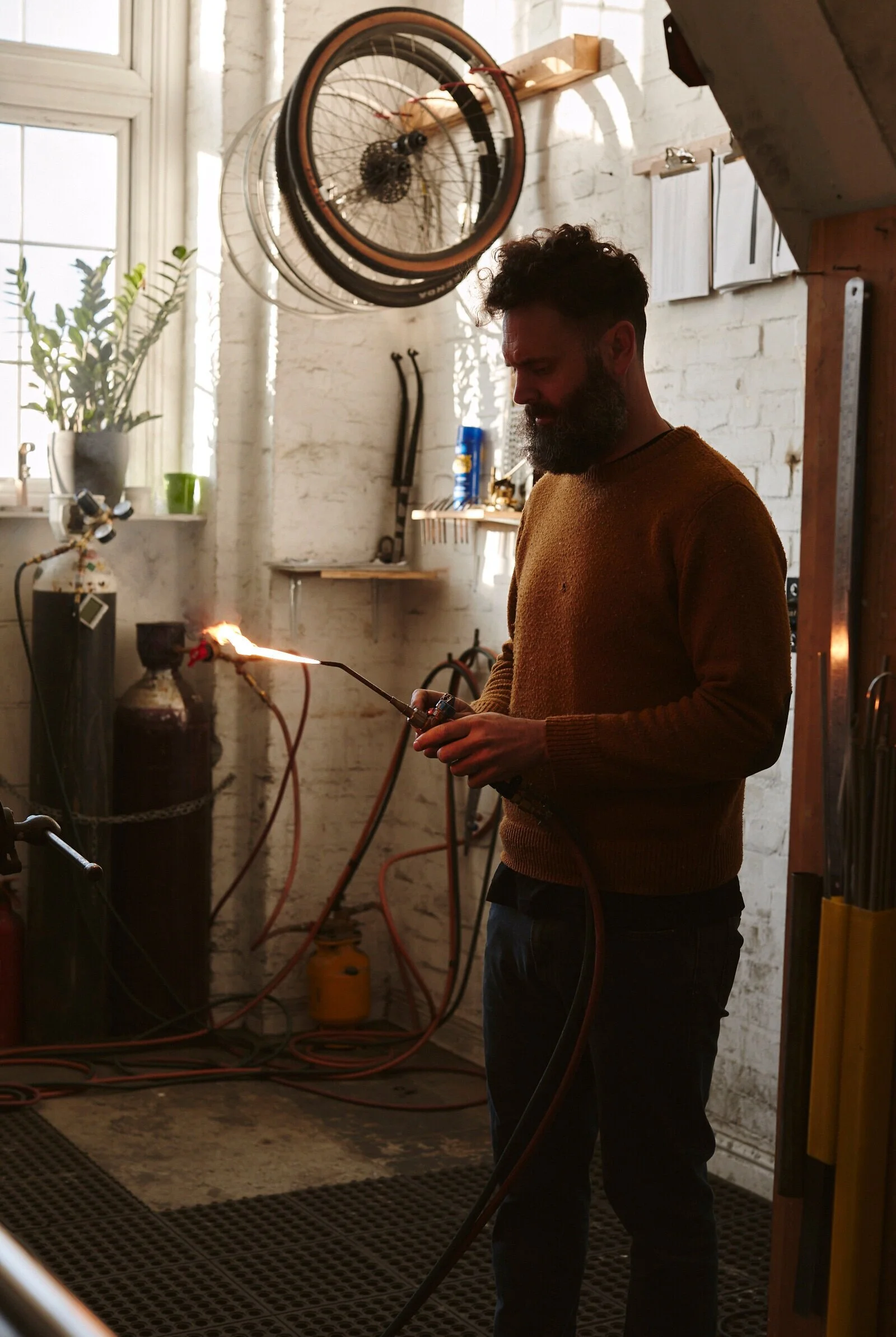 A man with a beard wearing a brown sweater is welding in a workshop, with a bicycle hanging on the wall above him.
