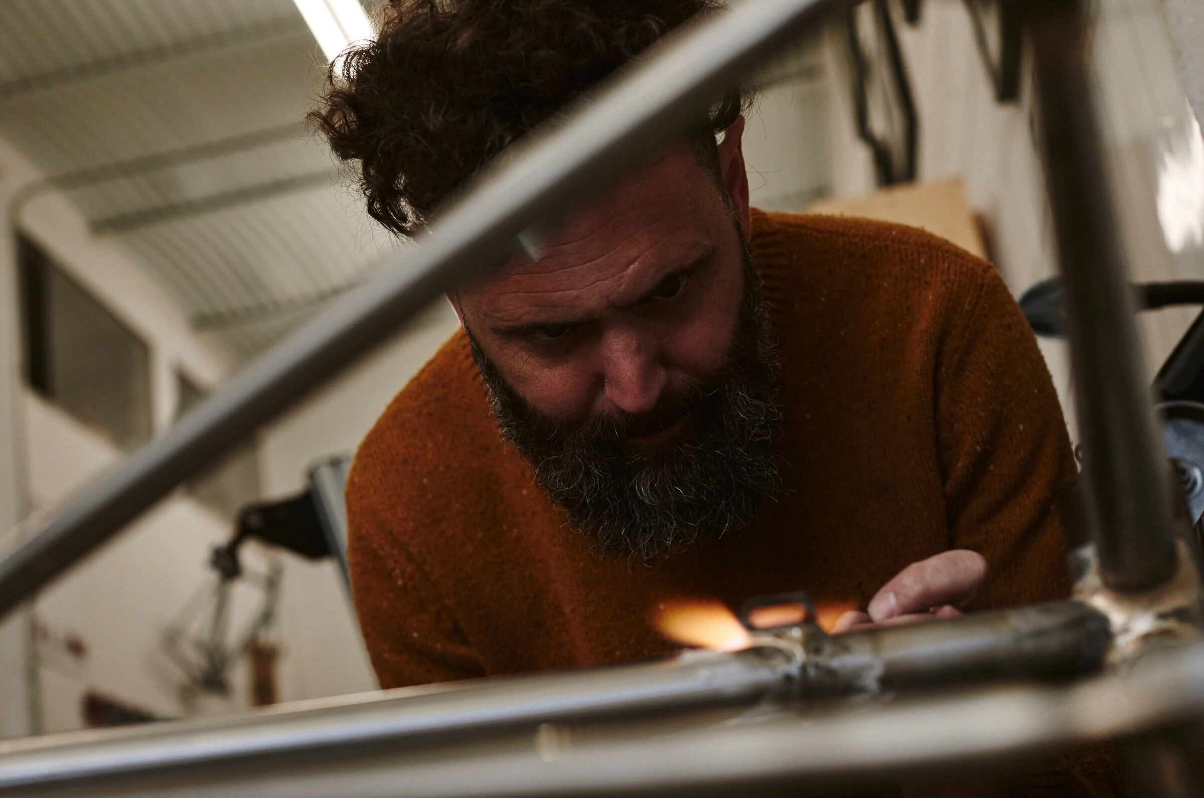A man with a beard and curly hair, wearing a brown sweater, is focused on welding metal at a workbench in a workshop.