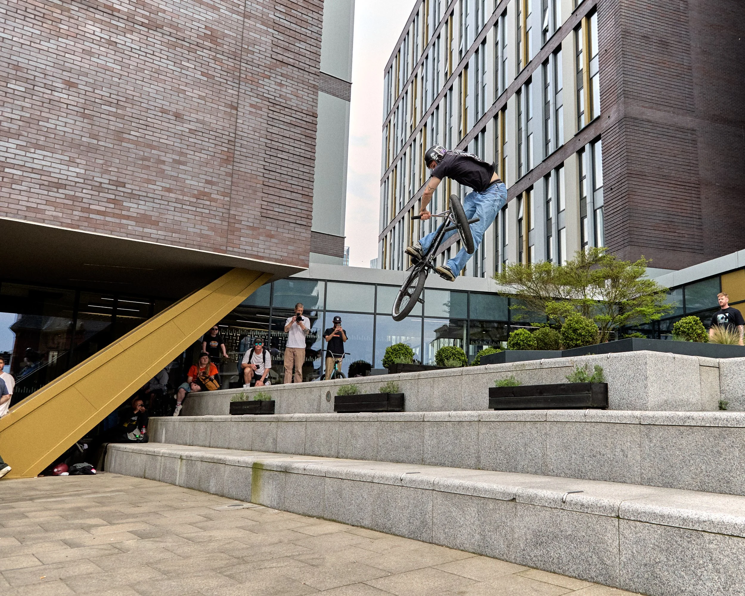 A BMX rider in mid-air performing a trick off a ledge in an urban setting with tall buildings and spectators watching.
