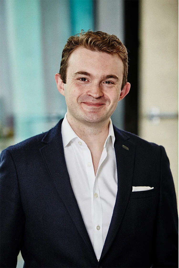 A young man with short brown hair wearing a dark suit jacket, white shirt, and pocket square, standing in an office or professional setting with a blurred background.