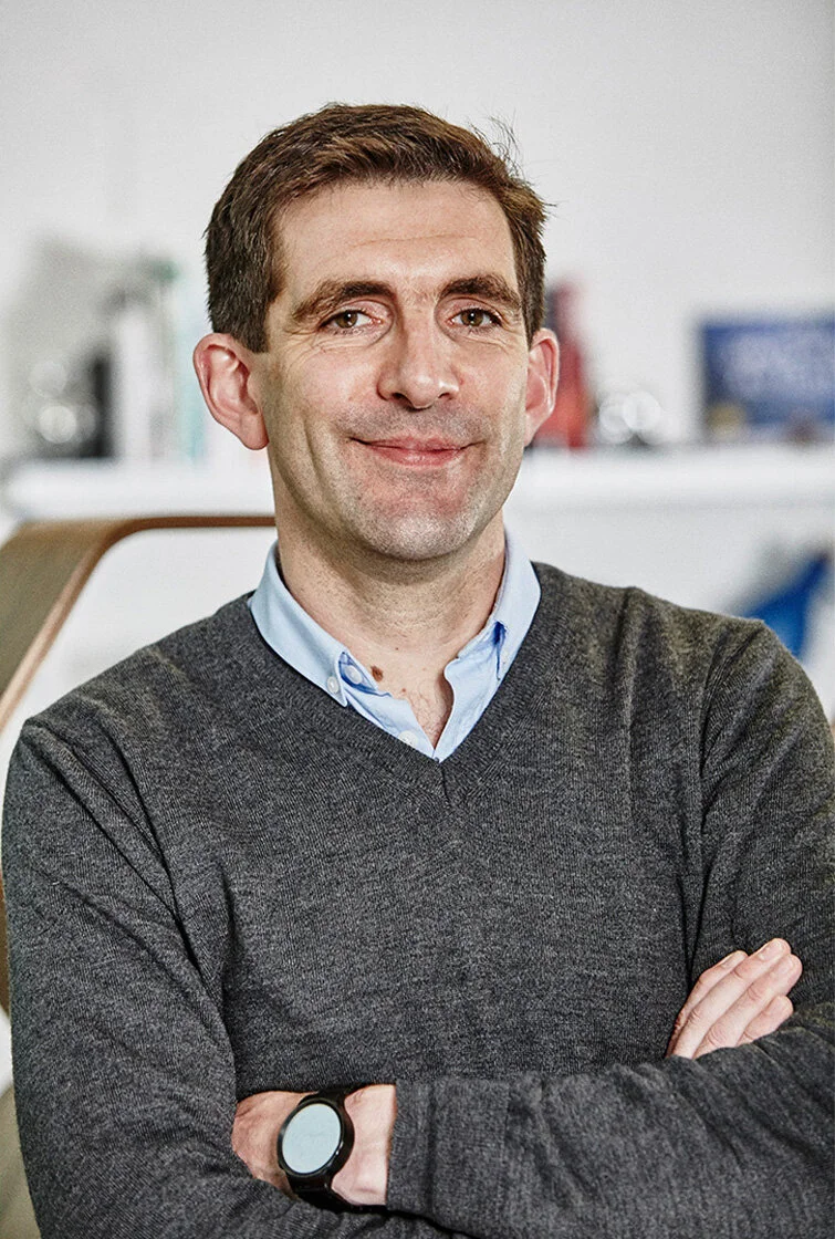 Portrait of a man with brown hair, wearing a dark gray sweater over a light blue collared shirt, standing with arms crossed in an office setting.