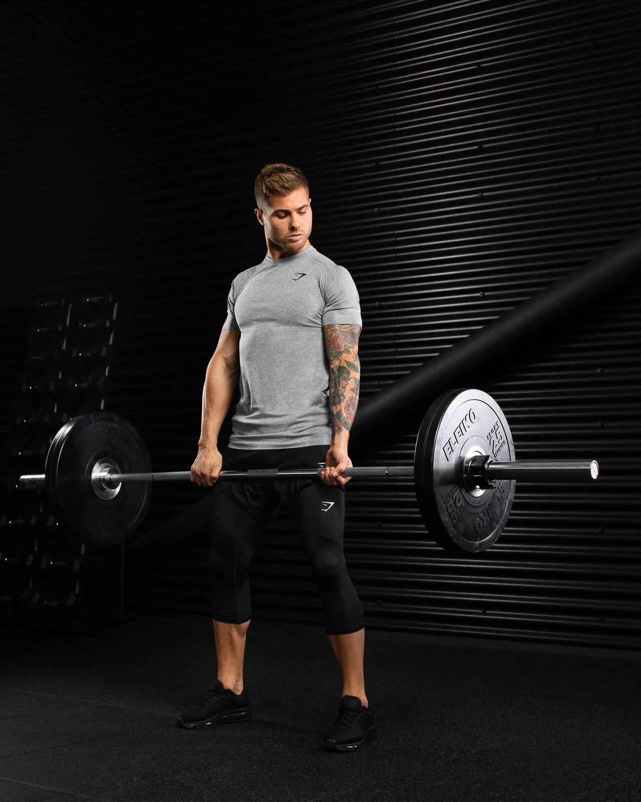 A man lifting a barbell loaded with weights in a gym, standing against a black wall with a textured surface.