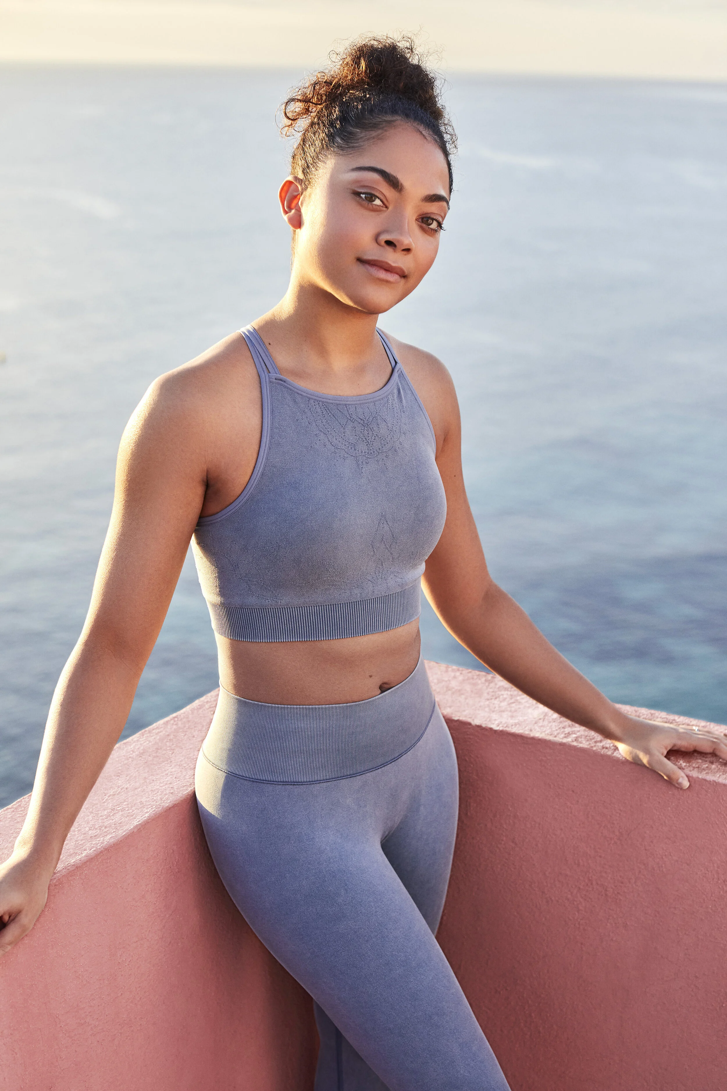Young woman in blue workout clothes standing by a pink wall near water, during sunset or sunrise.