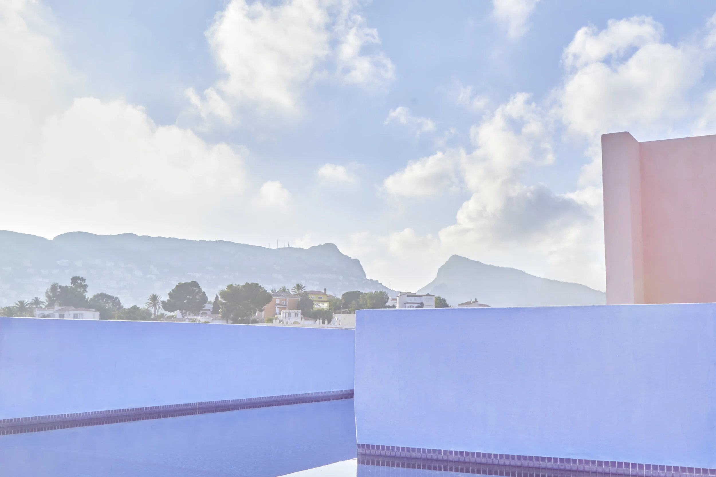 View of a swimming pool with a pastel pink and blue wall, distant mountains, a partly cloudy sky, and some trees and houses on the horizon.