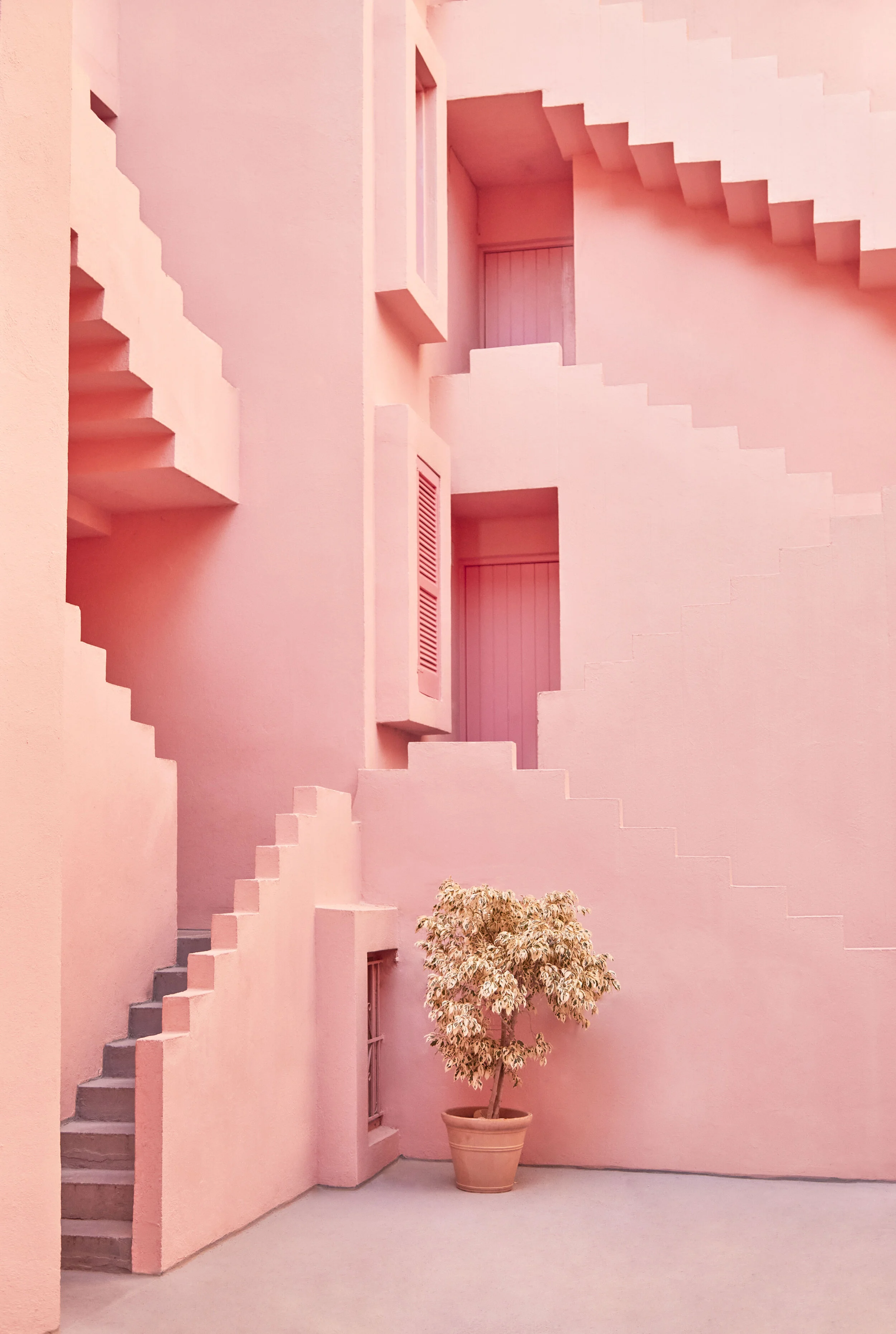 Pink building facade with geometric staircases and small windows, and a potted plant in the courtyard.