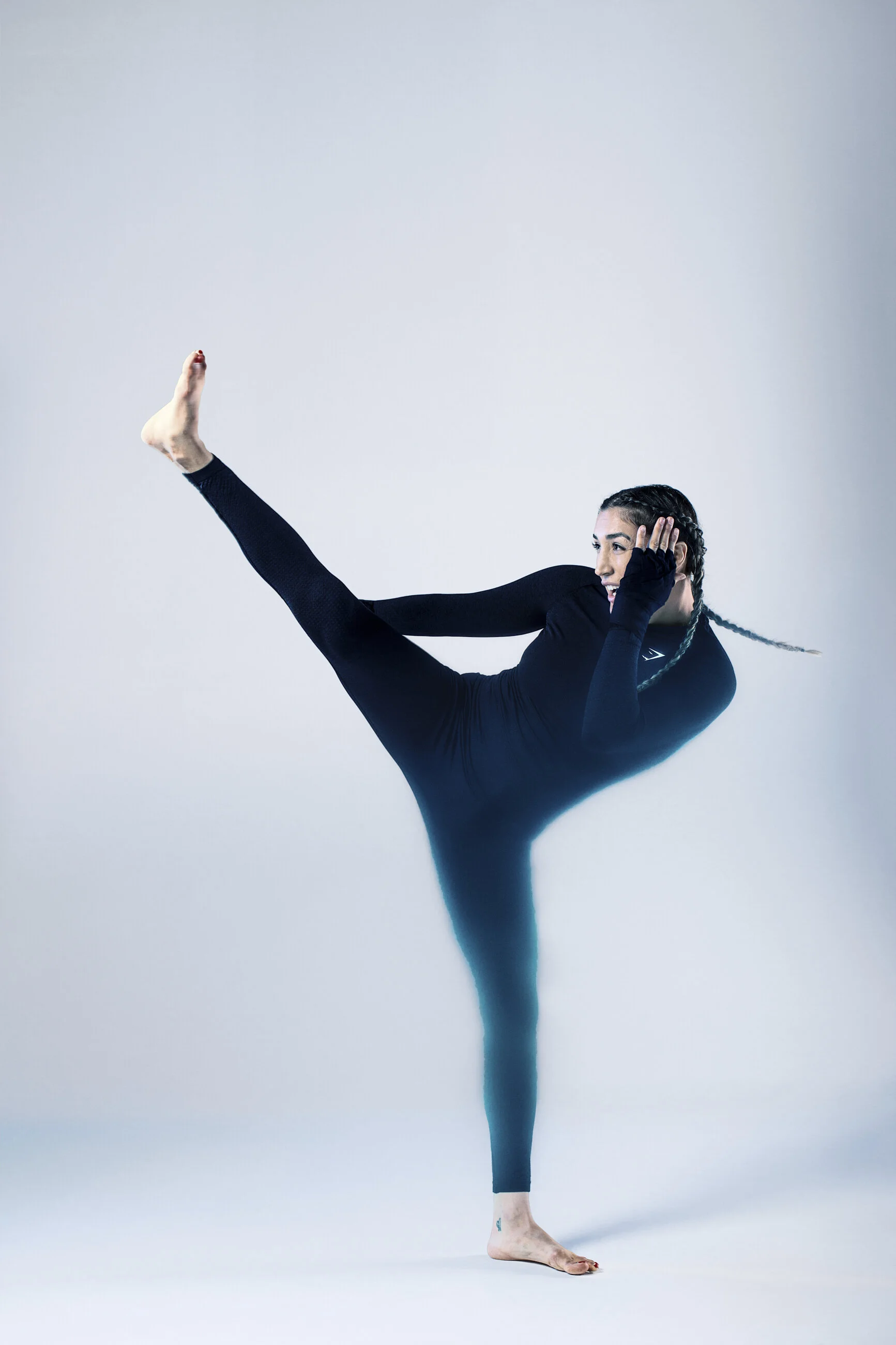 A woman in black activewear performing a yoga pose with one leg extended sideways and one hand on her head, against a plain light background.