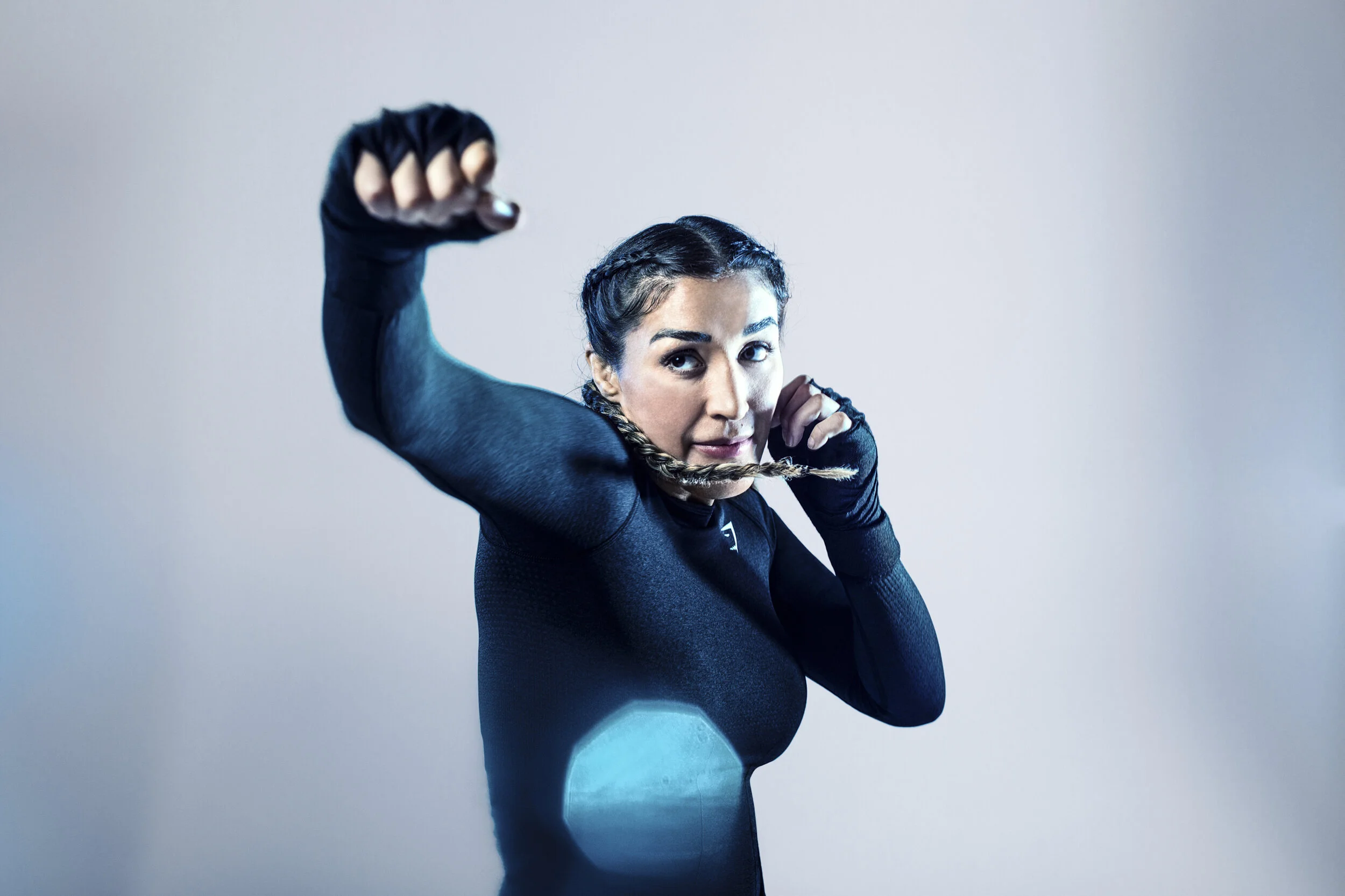 A woman in black athletic wear, with braided hair, flexes her arm and holds a braid in her mouth, smiling confidently.