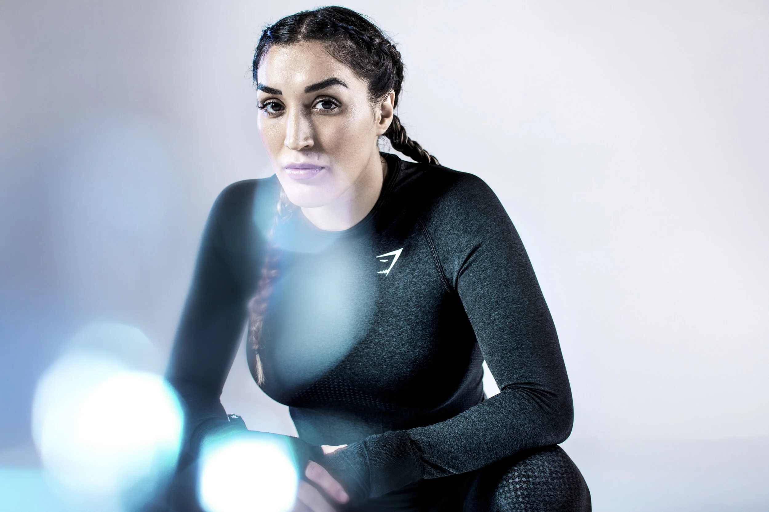 A woman with braided hair wearing a black athletic top, sitting against a plain light-colored background, looking at the camera with a slight smile.