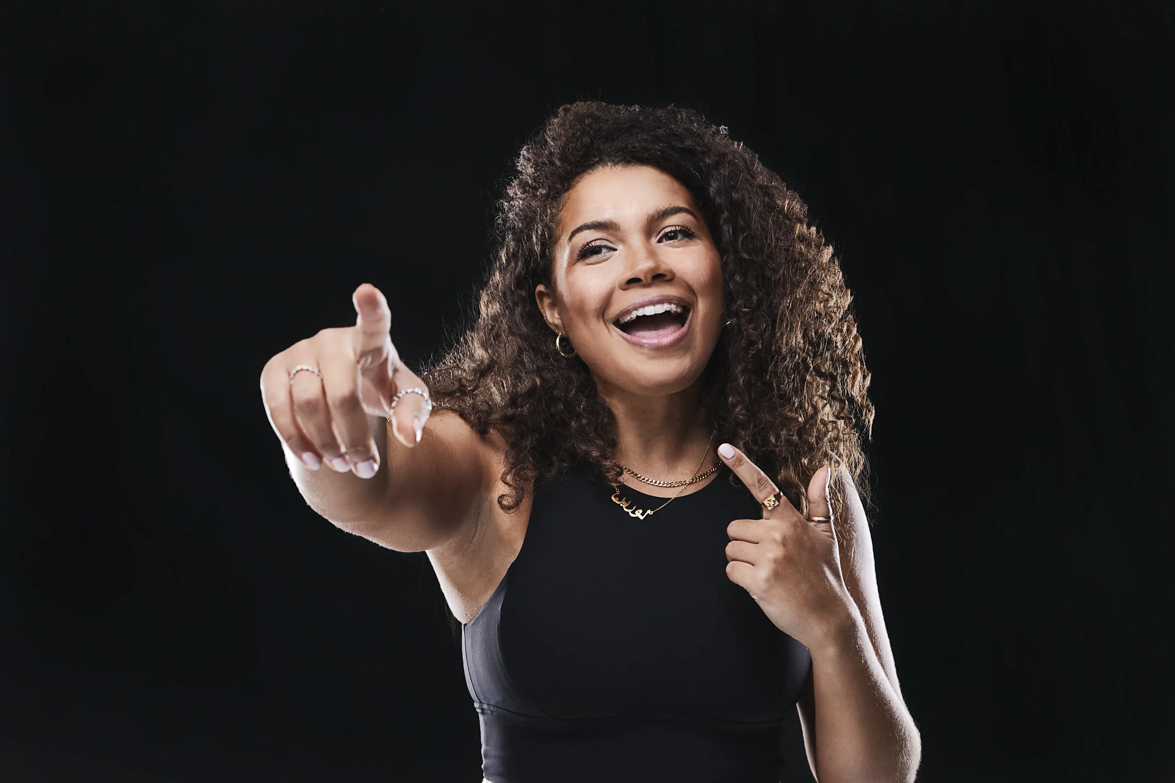 A woman with curly hair smiling, pointing forward, dressed in black, against a black background.