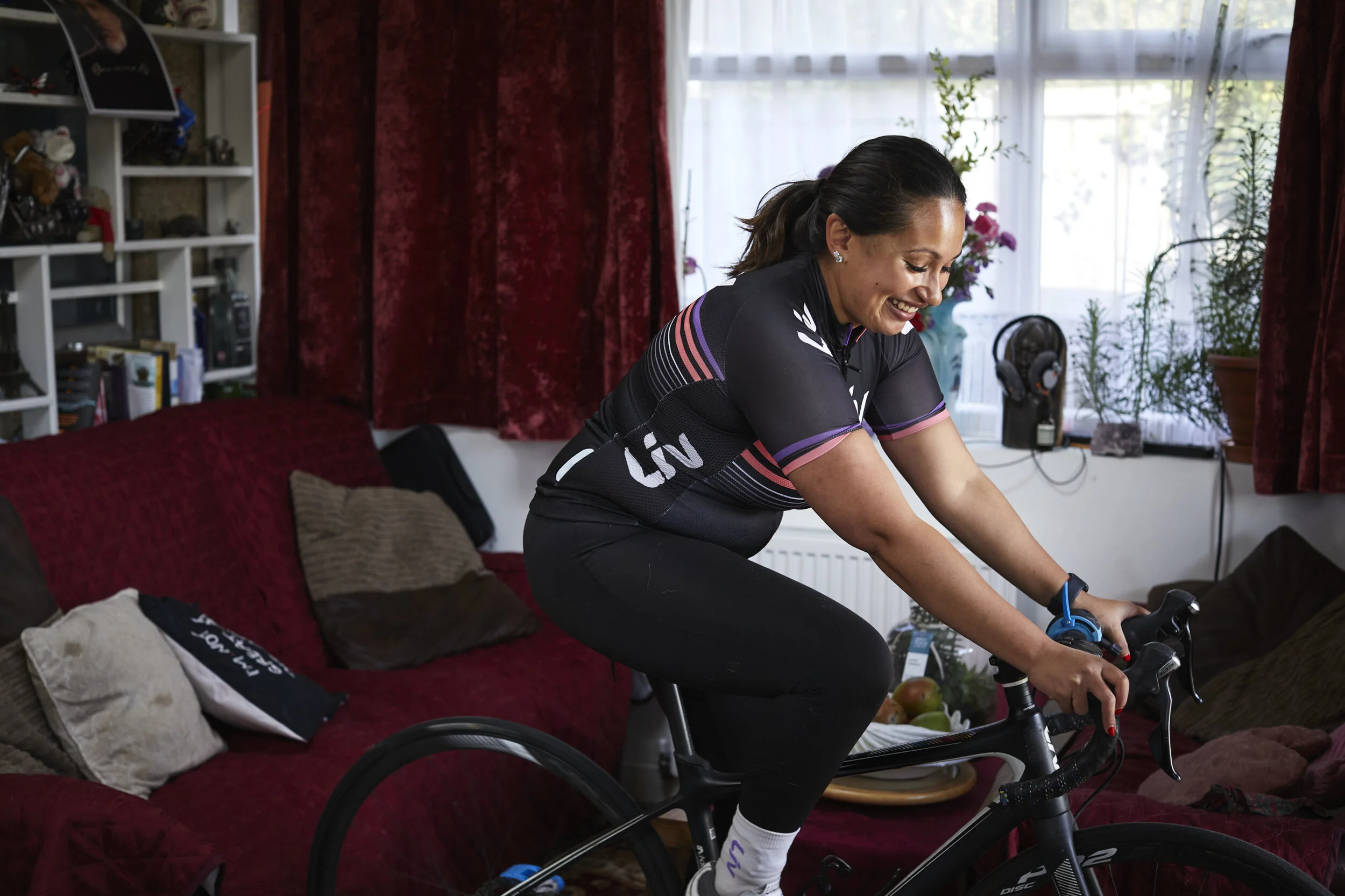 woman in sports cycling attire riding a stationary bike indoors, smiling with a living room background with red curtains, green plants, and shelves.