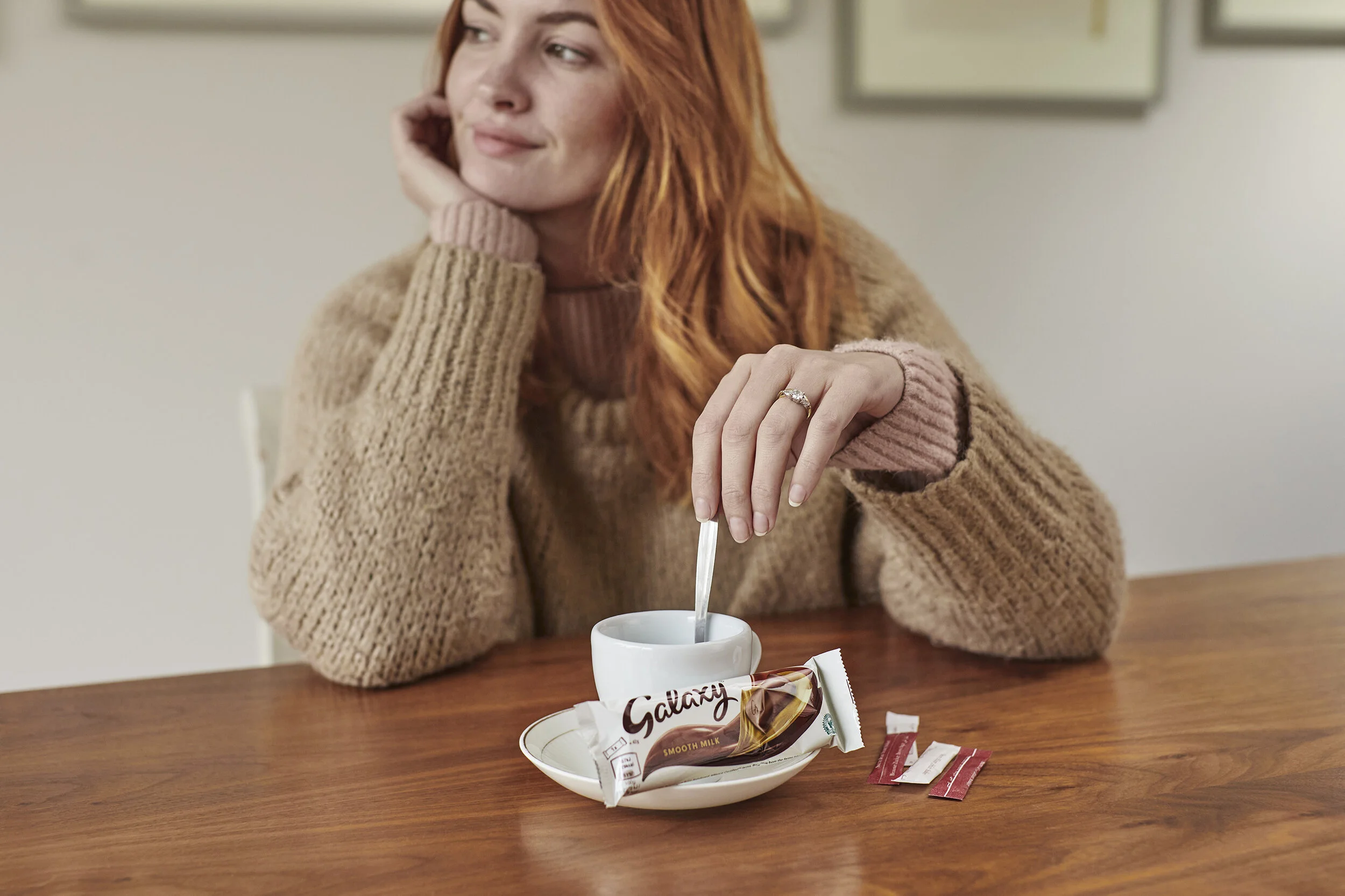A woman with red hair wearing a beige sweater sitting at a wooden table with a white mug, a spoon, and a Galaxy smooth milk chocolate bar.
