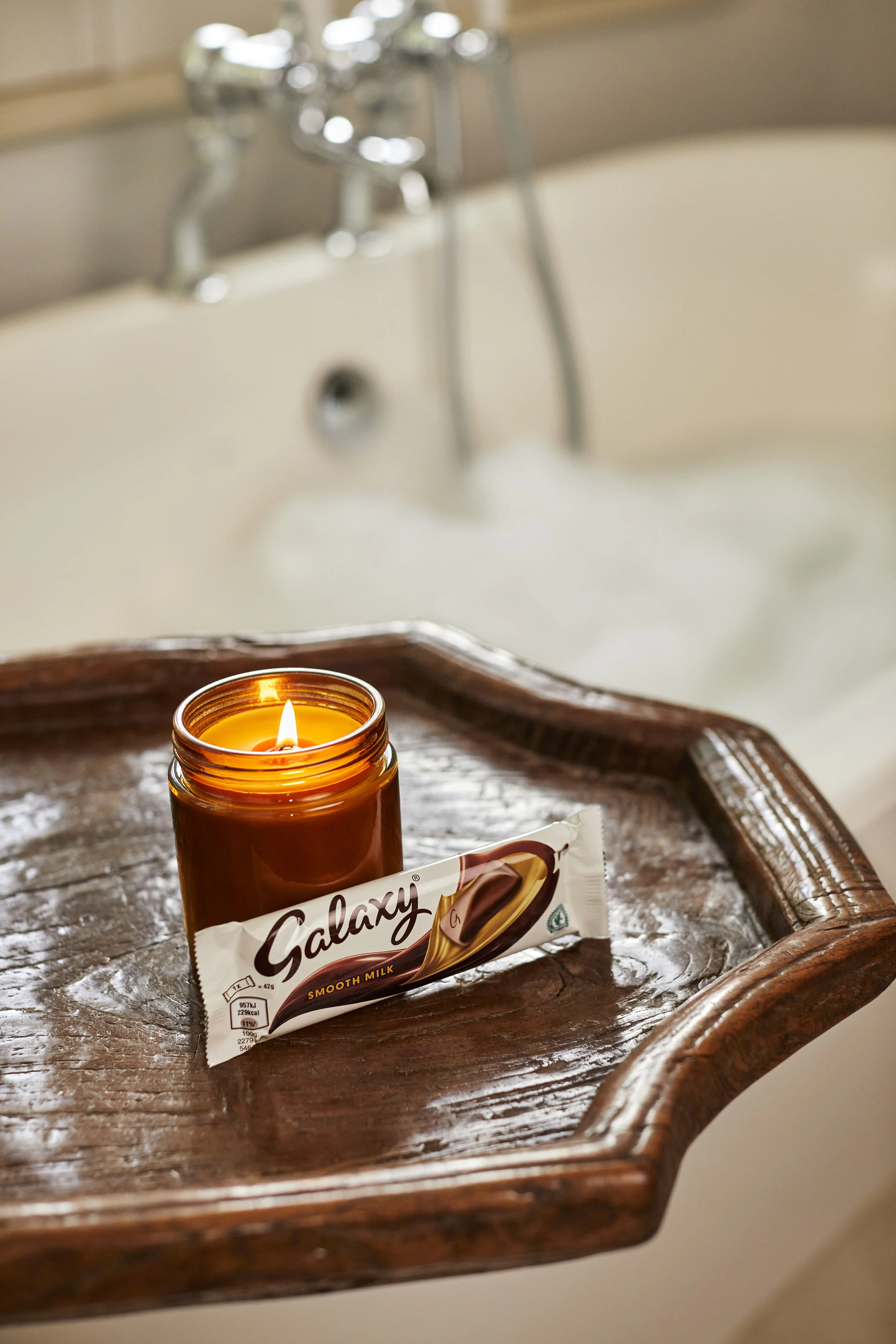 A lit candle and a Galaxy chocolate bar on a wooden tray next to a bathtub filled with water and soap suds.