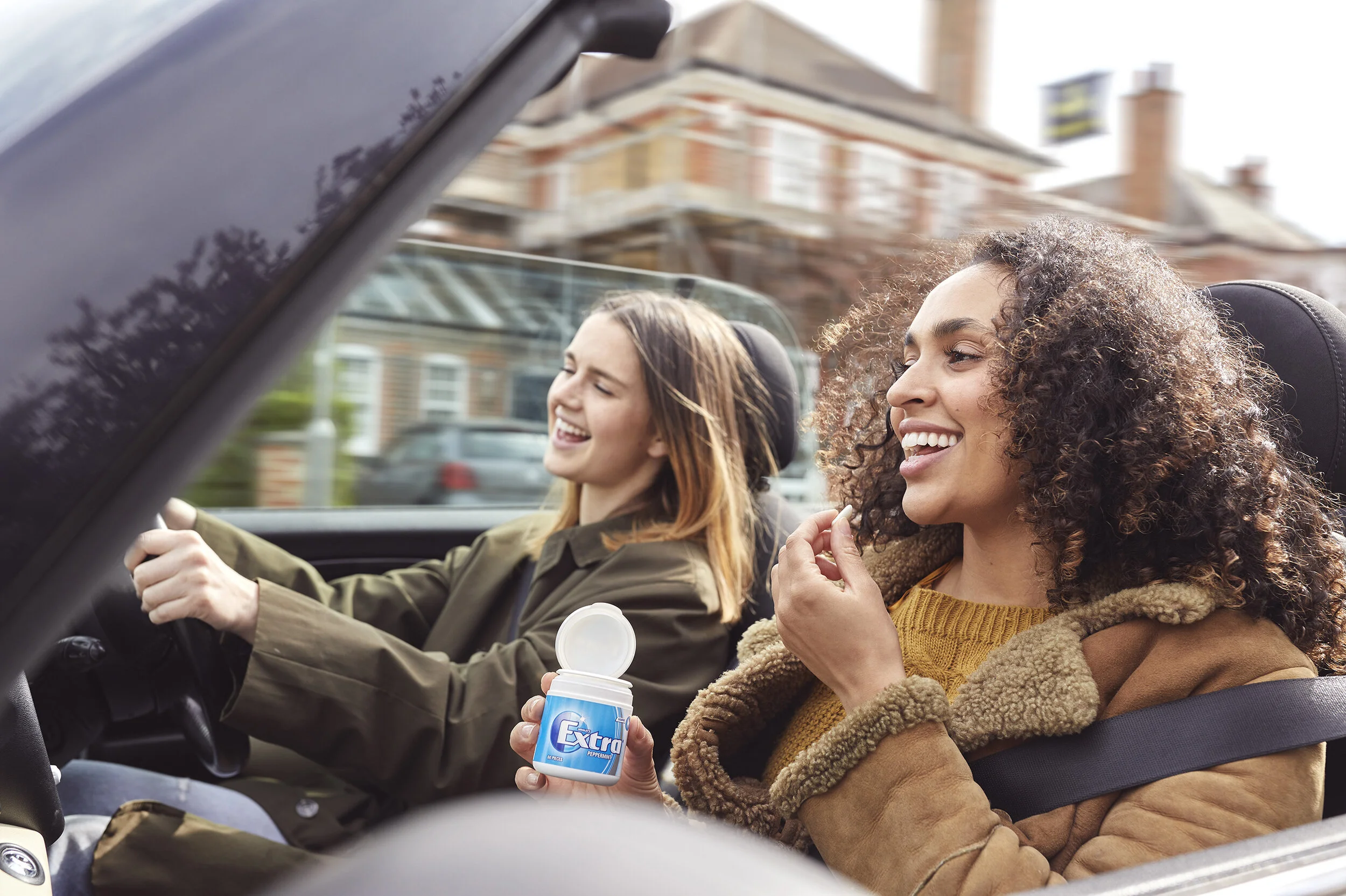 Two women are sitting in a convertible car, enjoying themselves. The woman in the passenger seat, with curly hair and wearing a tan coat, holds a container of Extra chewing gum and is smiling. The woman driving, with straight hair and wearing an oliv