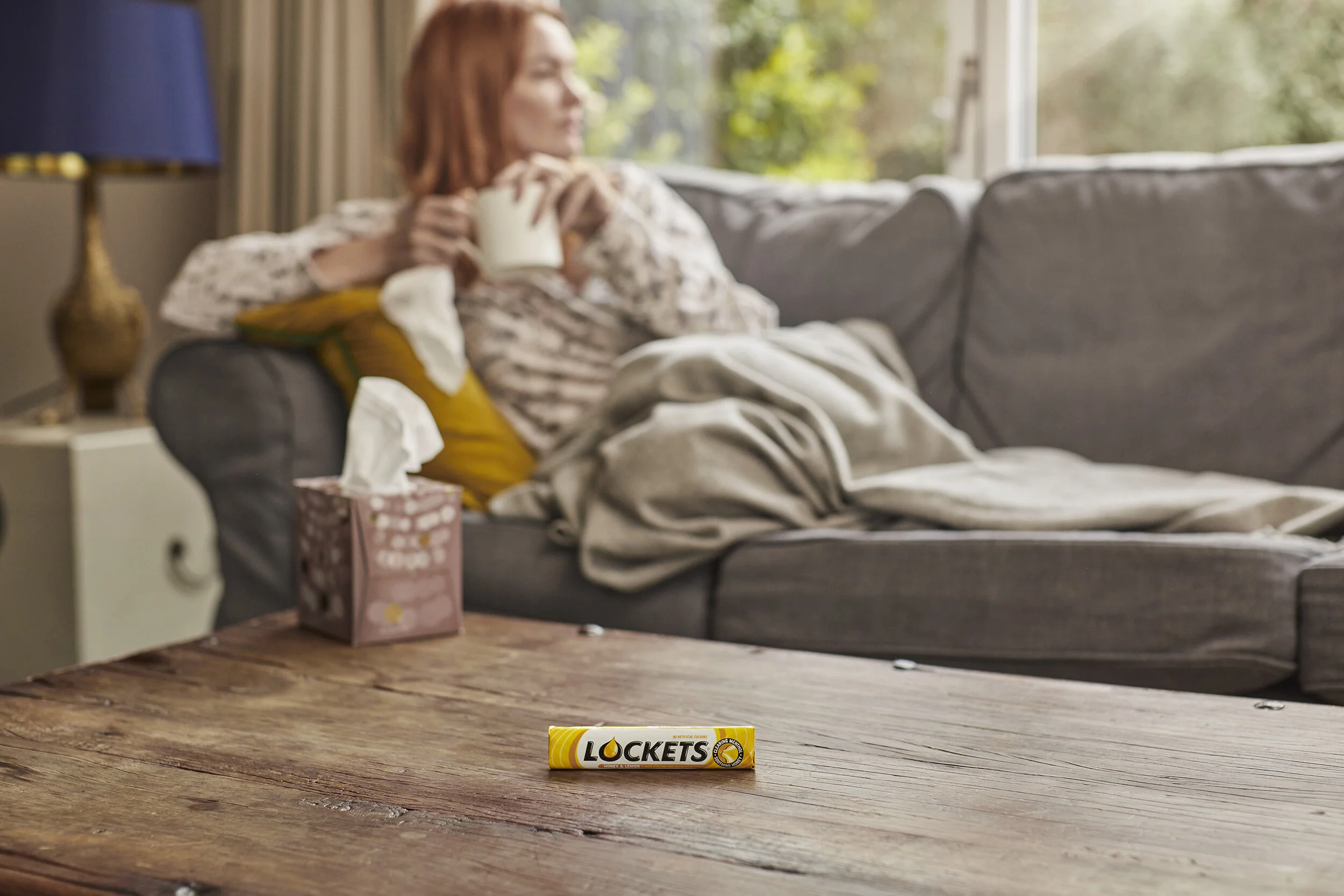 A woman sitting on a gray sofa holding a white mug, with a tissue box and a pack of Locket's tissues on a wooden table in front of her, and a window with greenery outside in the background.