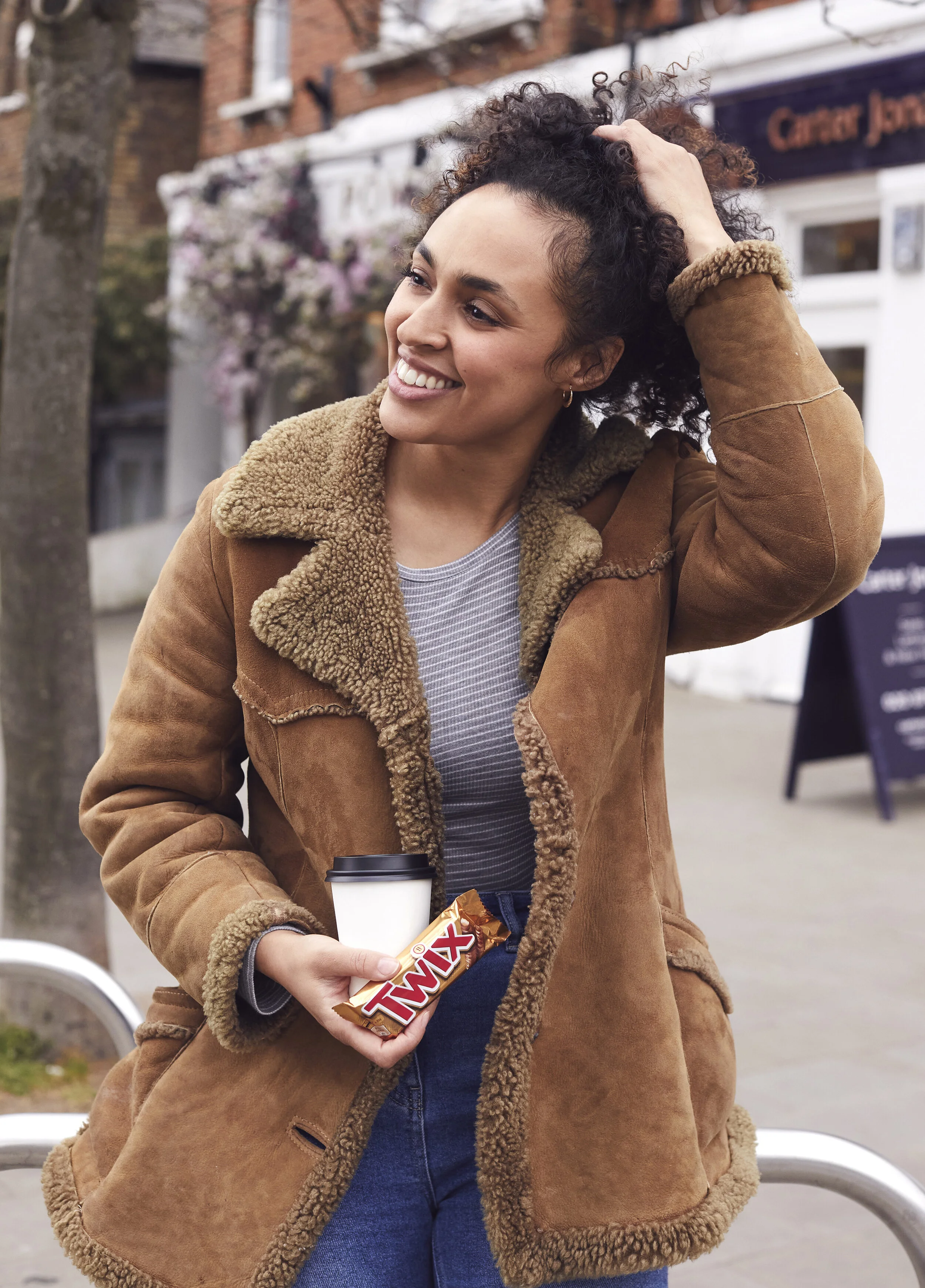 A woman with curly hair, smiling, holding a coffee cup and a Twix candy bar, standing outside on a sidewalk near a tree and a building.
