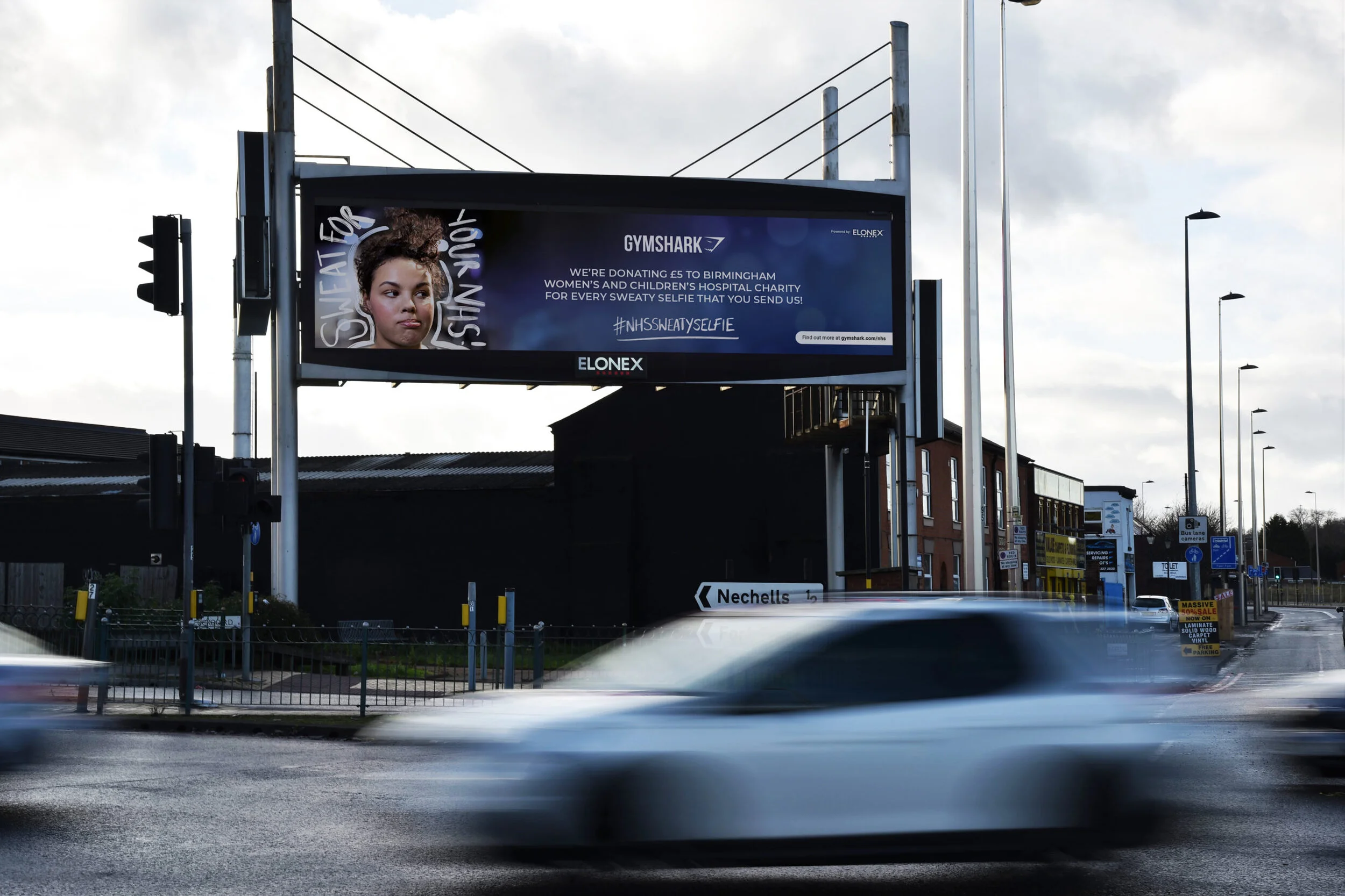 City street with moving cars and a billboard advertising Gymshark, donations for Birmingham women's and children's hospital, and a hashtag about sweat and selfie.
