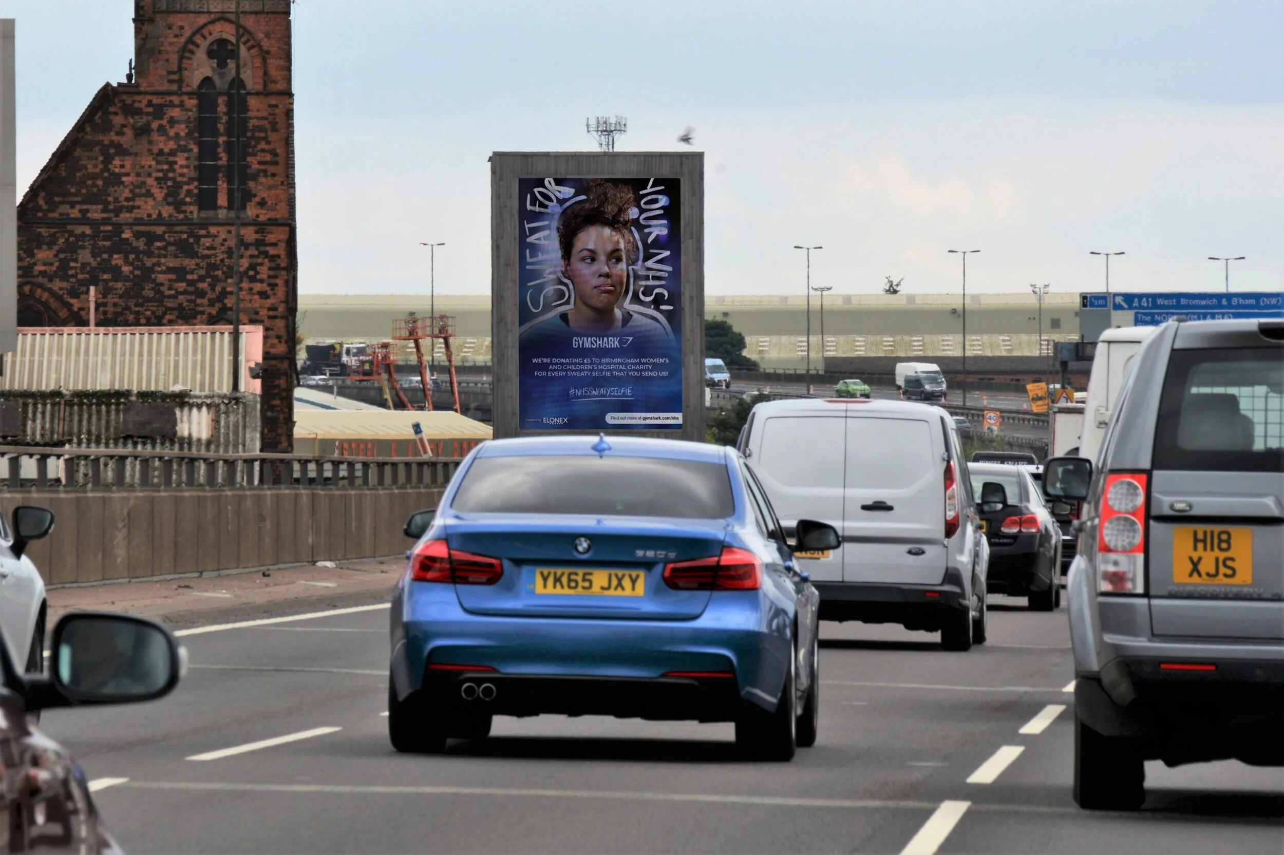 A busy road with multiple cars including a blue BMW, a white van, and other vehicles, with a billboard advertising Gymshark and a church in the background.