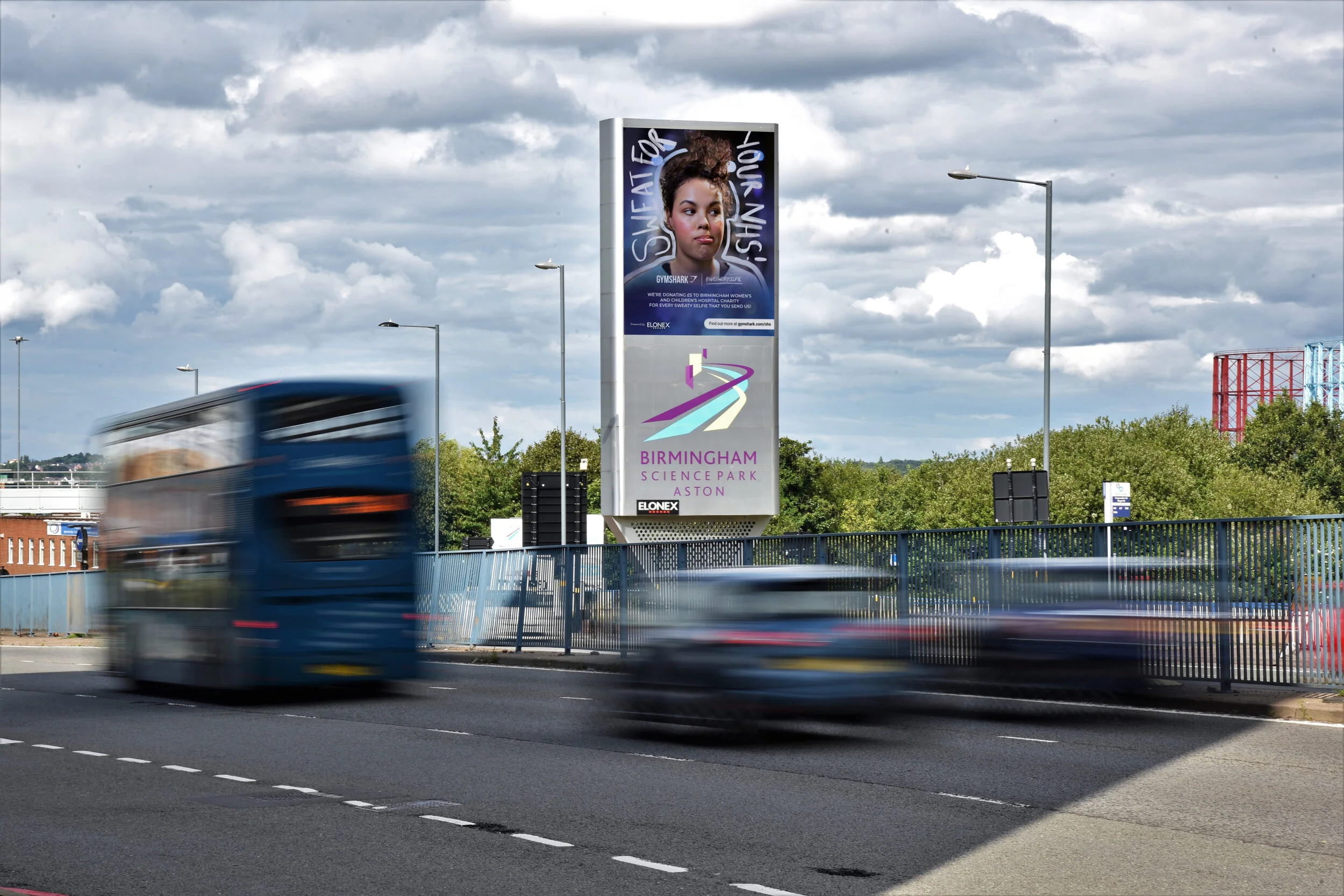 A large billboard on a city street with a woman’s portrait and text, and below it the Birmingham Science Park Aston logo. Blurred cars are passing on the road in front of the billboard under a cloudy sky.