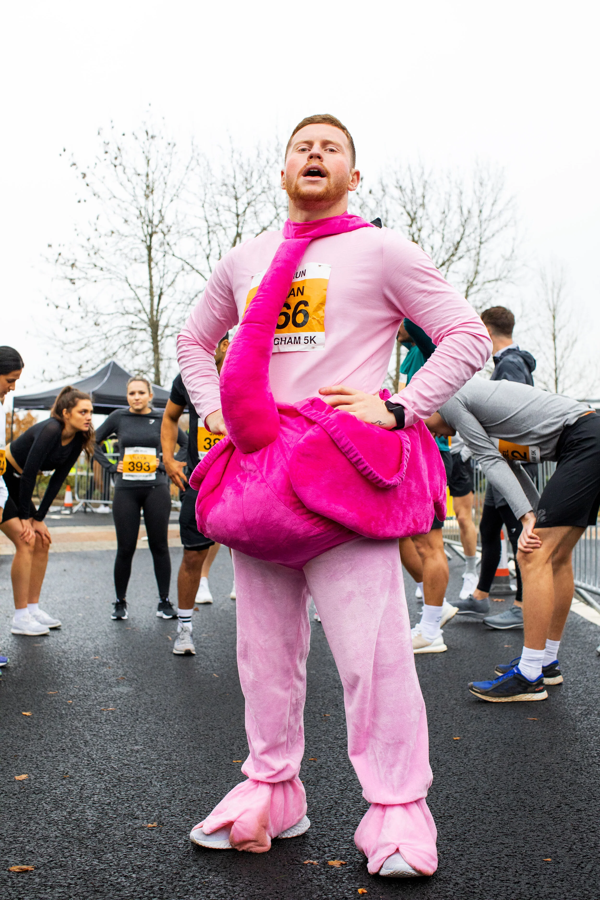 A man dressed in a pink flamingo costume participating in a running race, with other runners in athletic gear around him at the starting line.