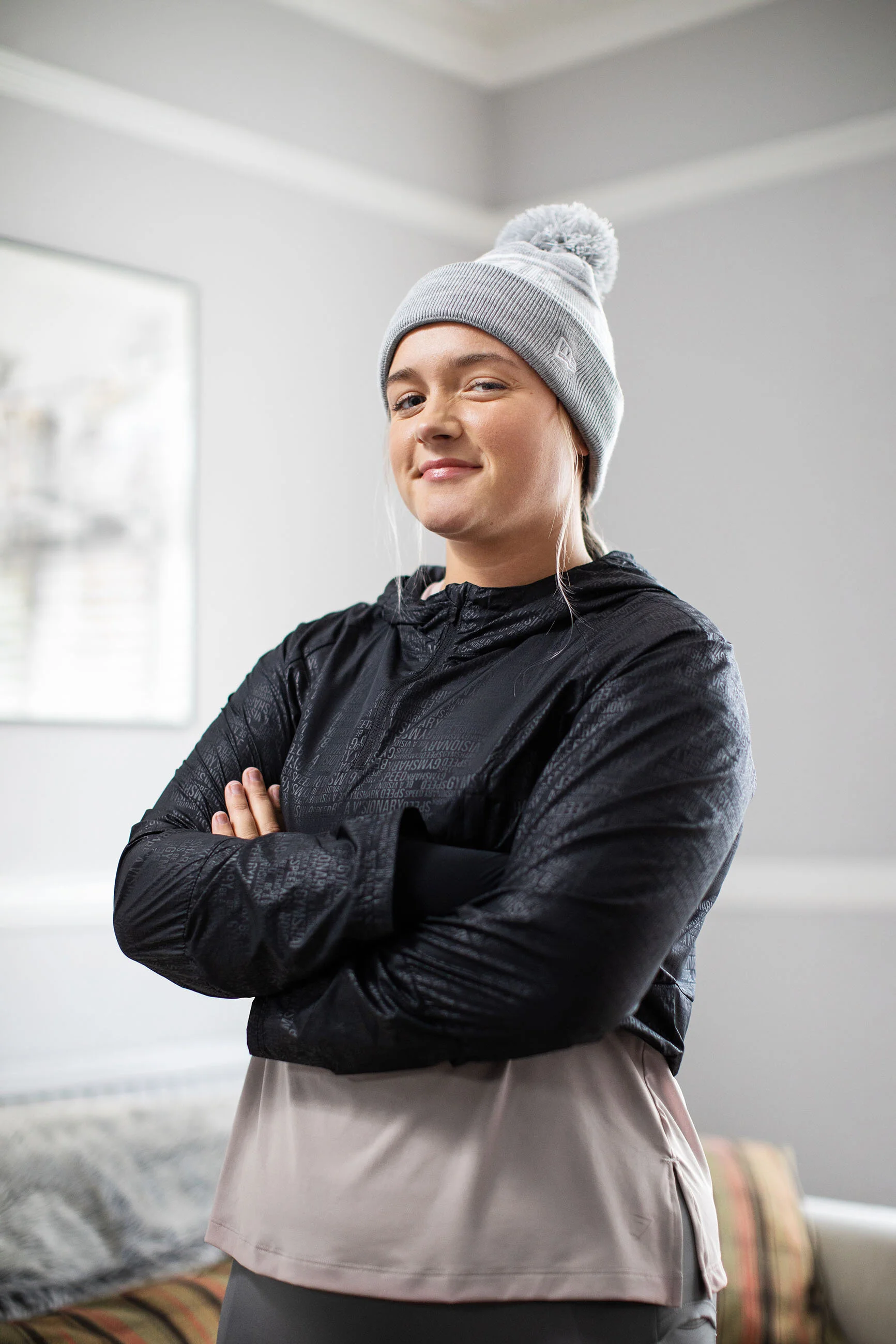 A young woman in athletic clothing, including a gray beanie and black jacket, standing indoors with her arms crossed and smiling at the camera.