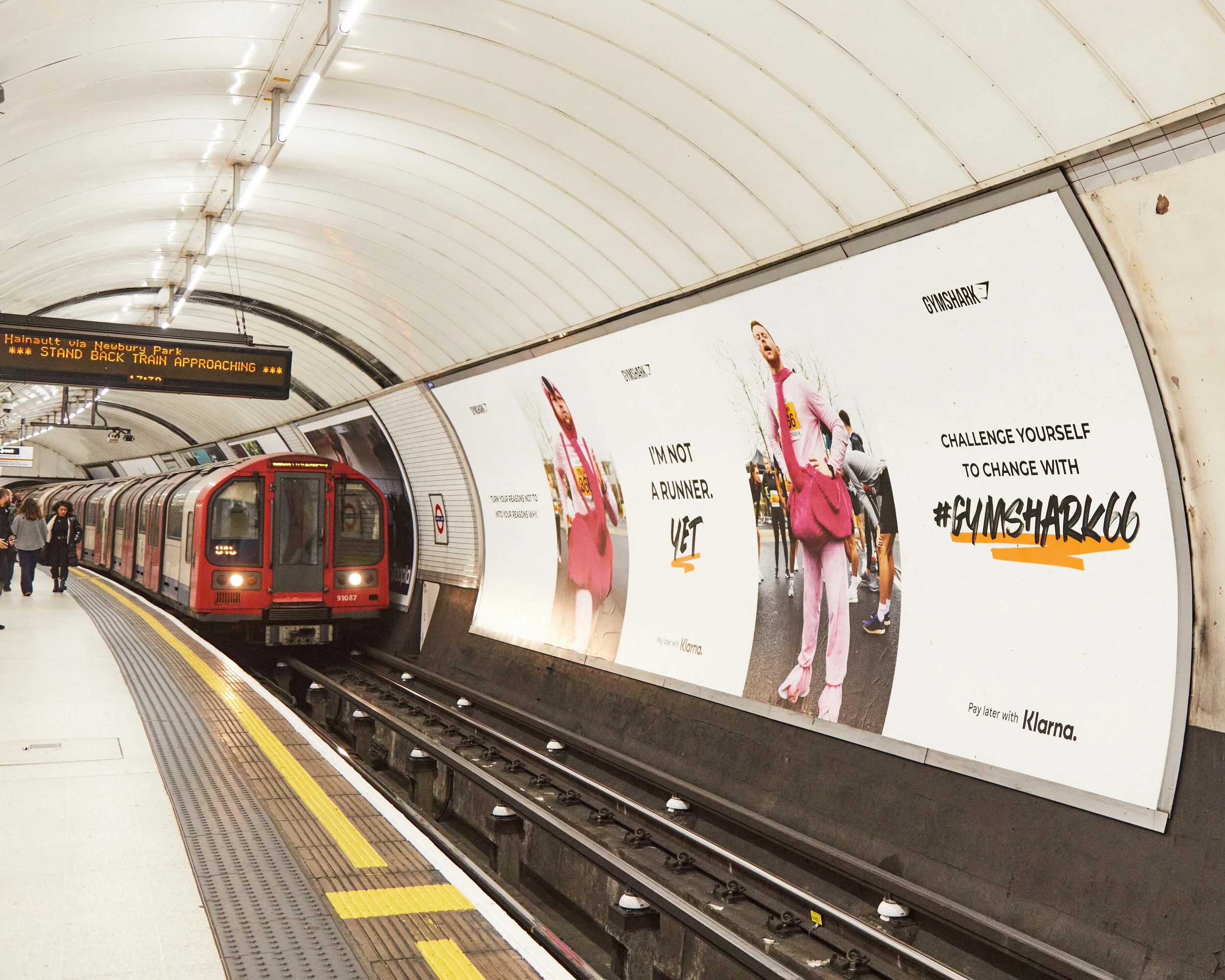 London Underground station platform with a red train stopped at the edge. There are passengers on the platform. A large advertisement on the wall shows a person in a pink outfit with a pink bag and an inspiring quote, along with the Gymshark logo and