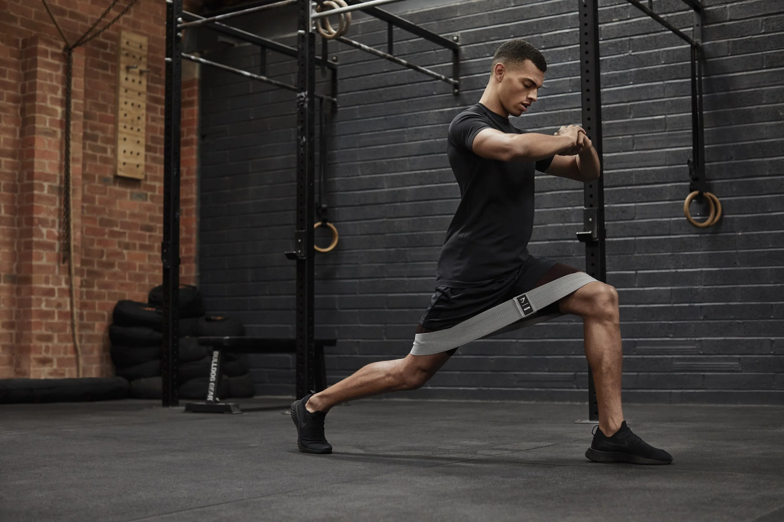 A man is performing a stretching exercise in a gym, with his hands clasped and pushed forward, one leg bent forward with a resistance band around his thigh, and the other leg extended back.