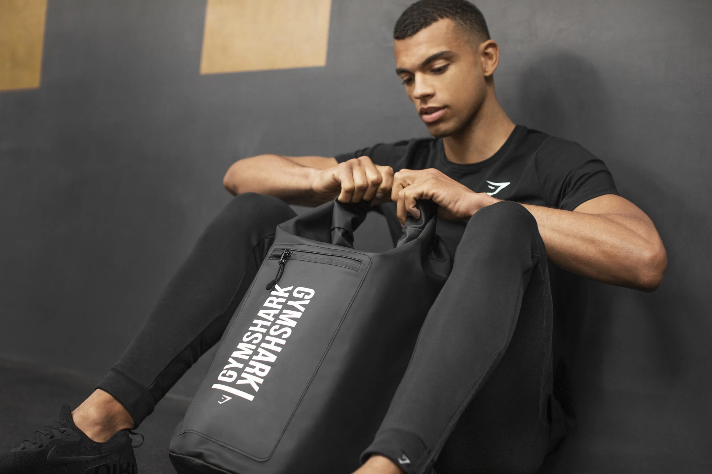 Young man sitting on the floor with a gym bag beside him, wearing athletic clothing and black sneakers.