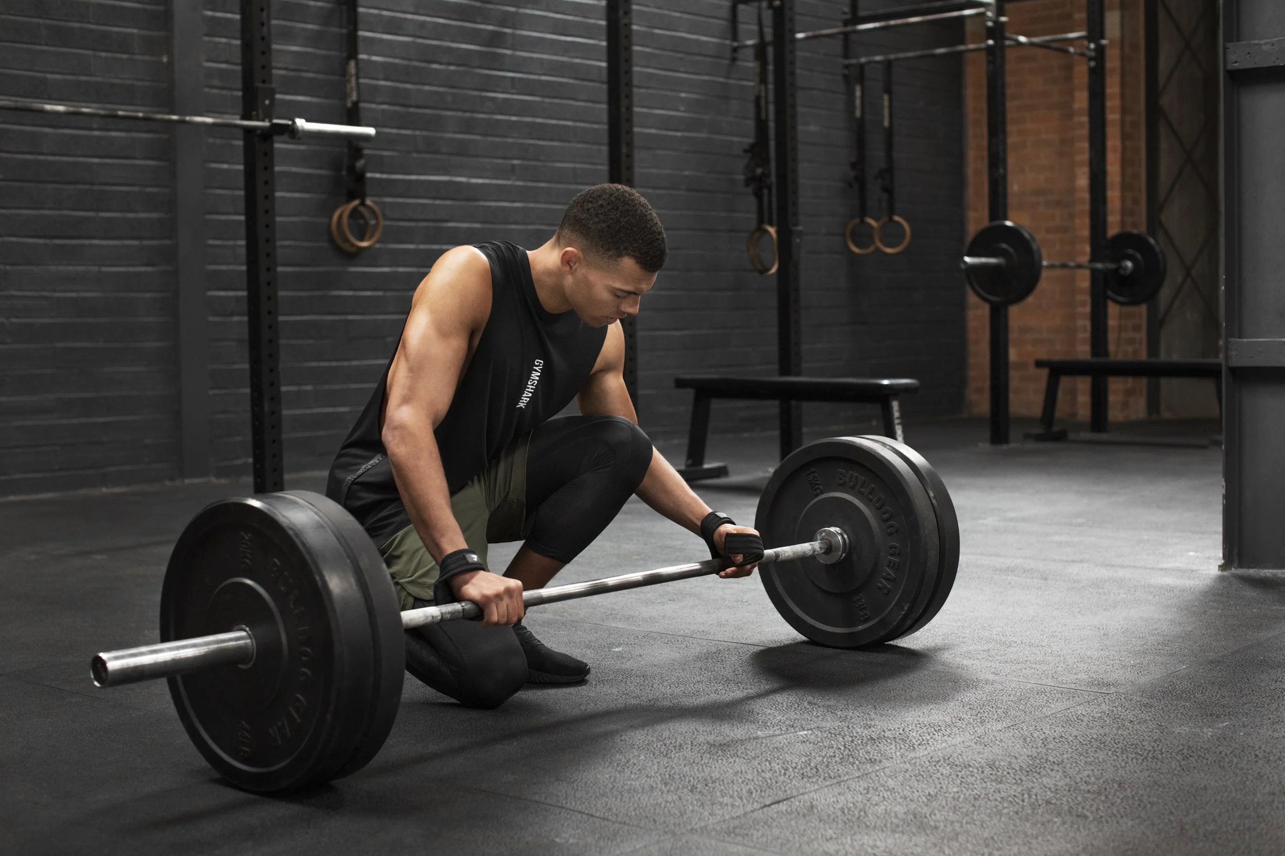 A man in workout gear kneeling on the gym floor, preparing to lift a barbell loaded with black weight plates.