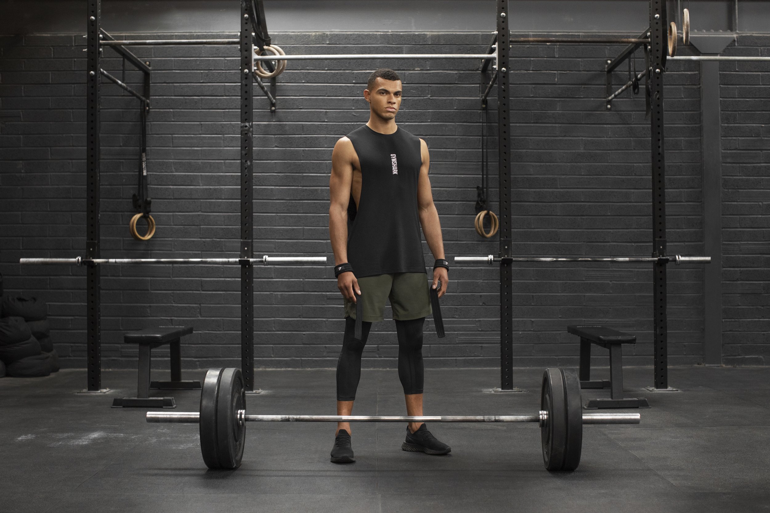 A young man standing in a gym with a barbell on the floor in front of him, black walls, and workout equipment in the background.