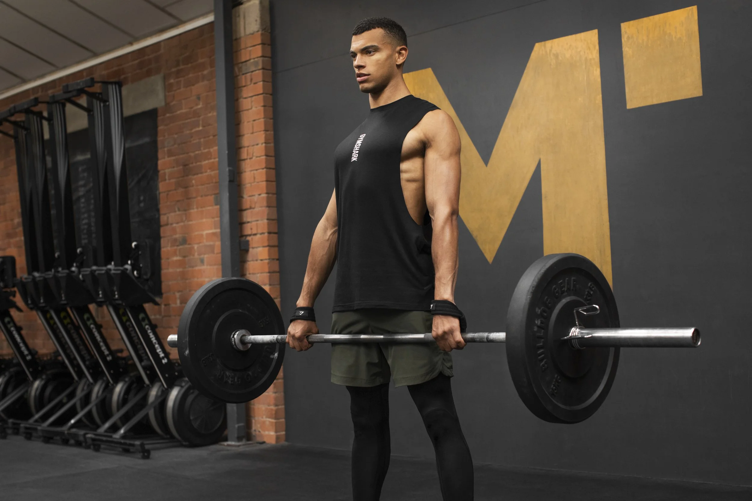 A man lifting a barbell in a gym, wearing a sleeveless black shirt and black leggings, with a brick wall and large yellow 'M' on the wall behind him.