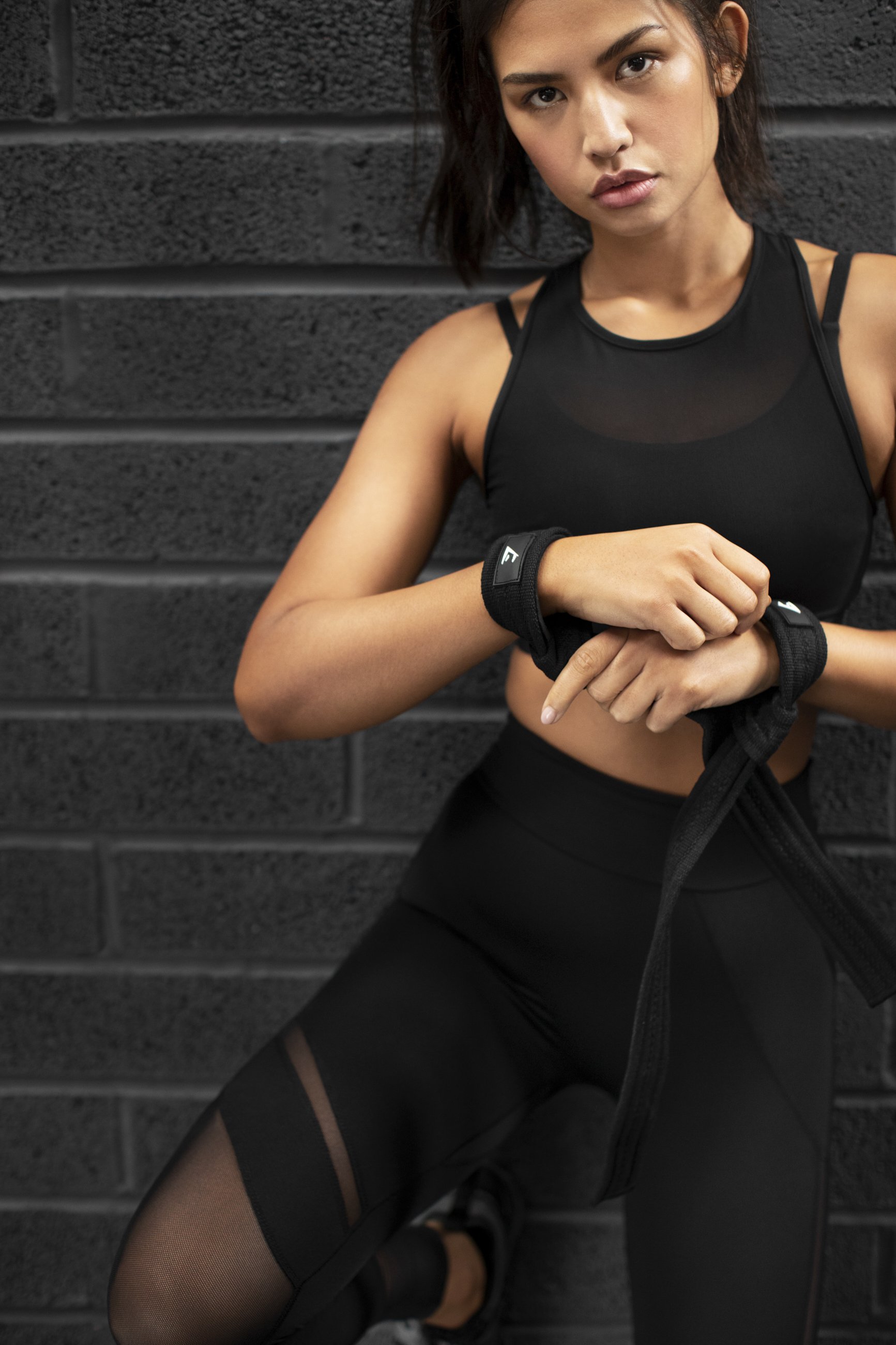 A fit woman in black athletic wear adjusting her wrist straps while standing against a black brick wall.