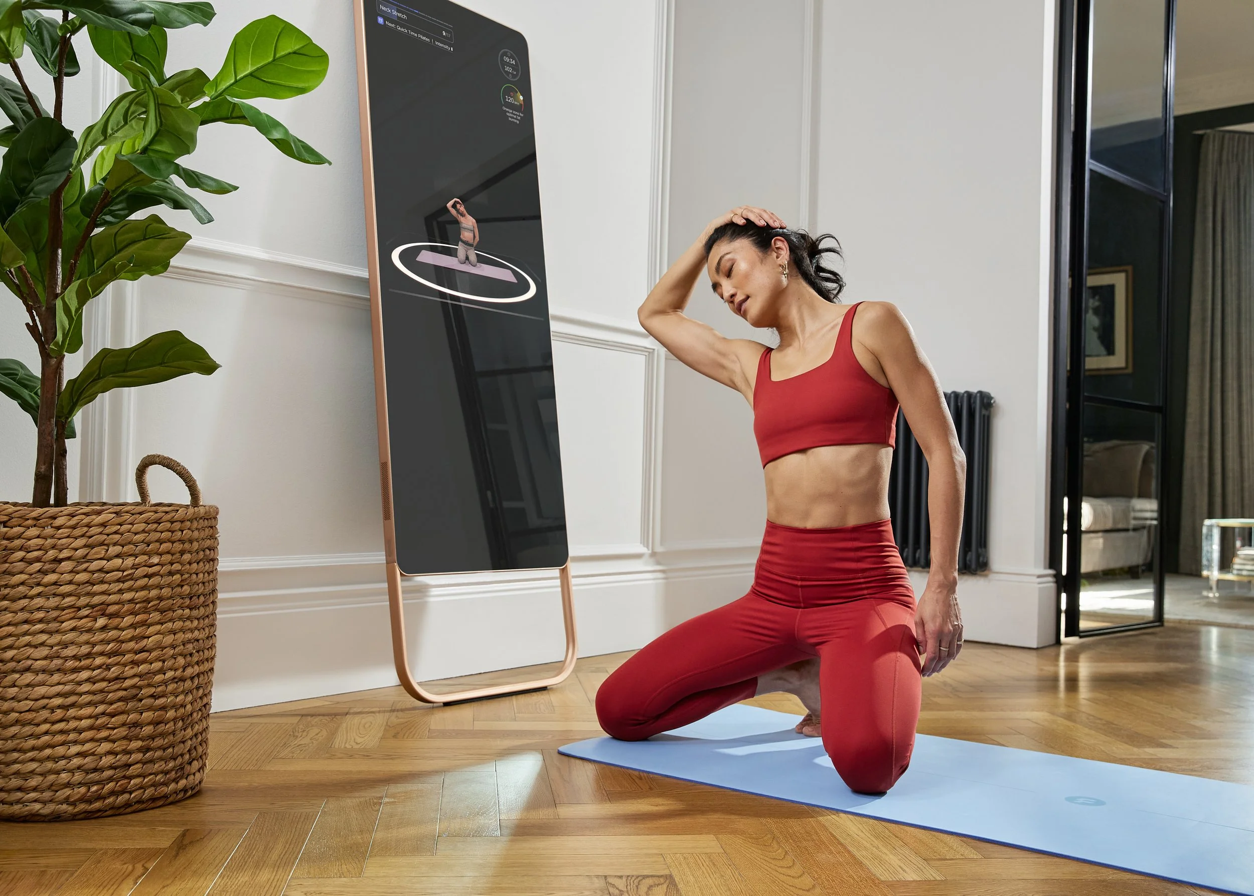A woman in red workout clothes kneeling on a yoga mat and holding her head in her hand in a room with hardwood floors, a large potted plant, and an electronic device displaying fitness data.