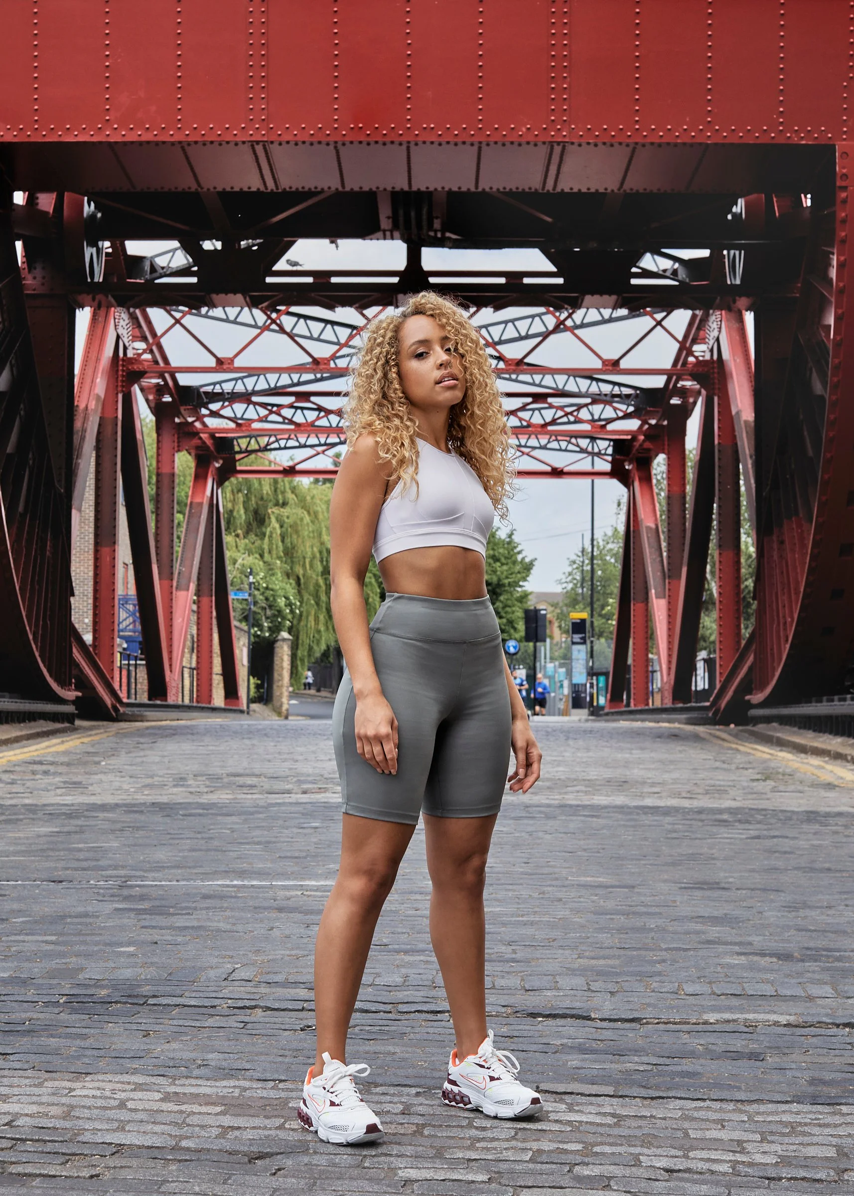 A woman in athletic wear standing on a cobblestone street under a red iron bridge.
